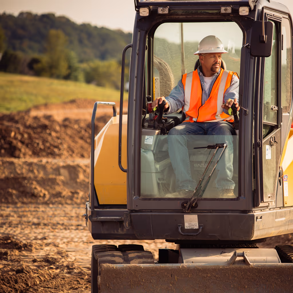 Construction worker in white hard hat and orange safety vest operating excavator at a dirt site with green trees in the background.