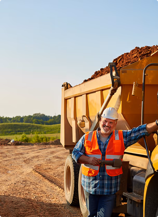 Construction worker wearing a white hard hat and orange safety vest, smiling and standing beside a large yellow dump truck loaded with dirt on a construction site.
