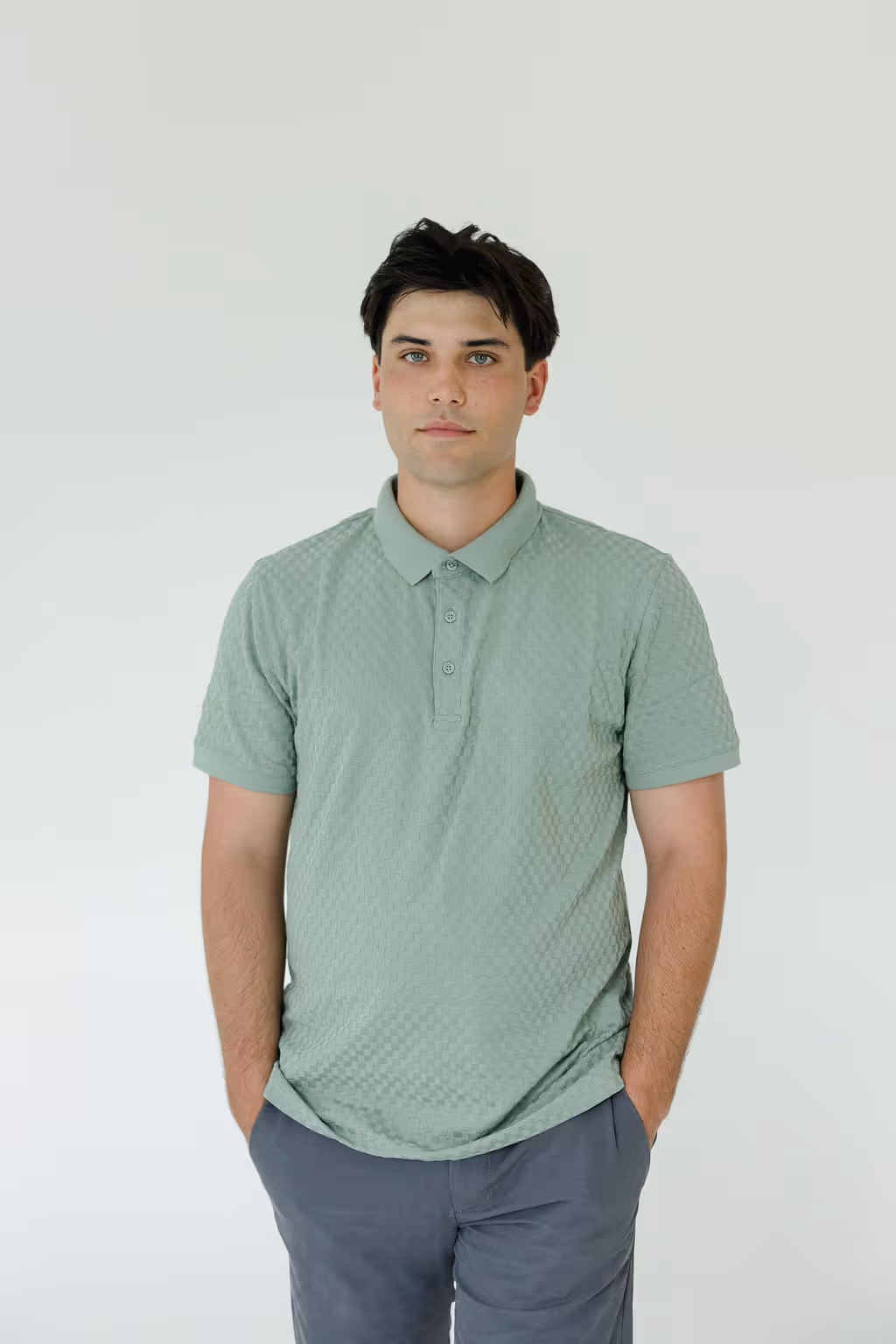 Young man with dark hair wearing a light green textured polo shirt and gray pants standing with hands in pockets against a plain light background.