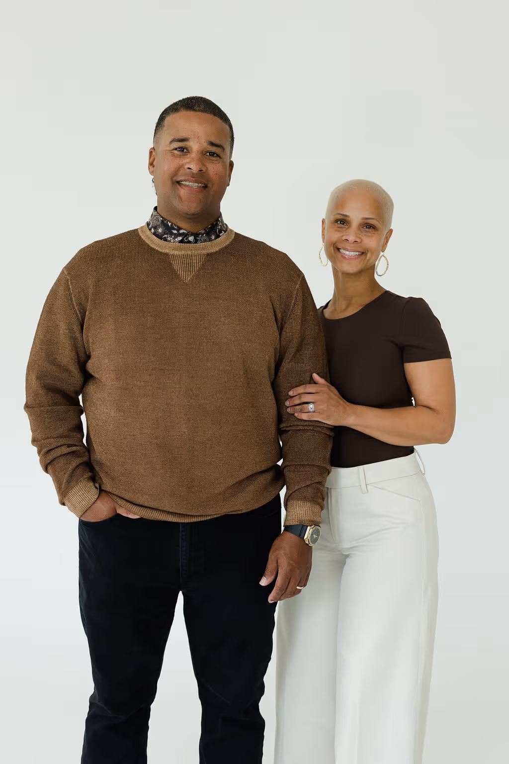 Smiling man in a brown sweater and woman in a black top and white pants standing close together against a light background.