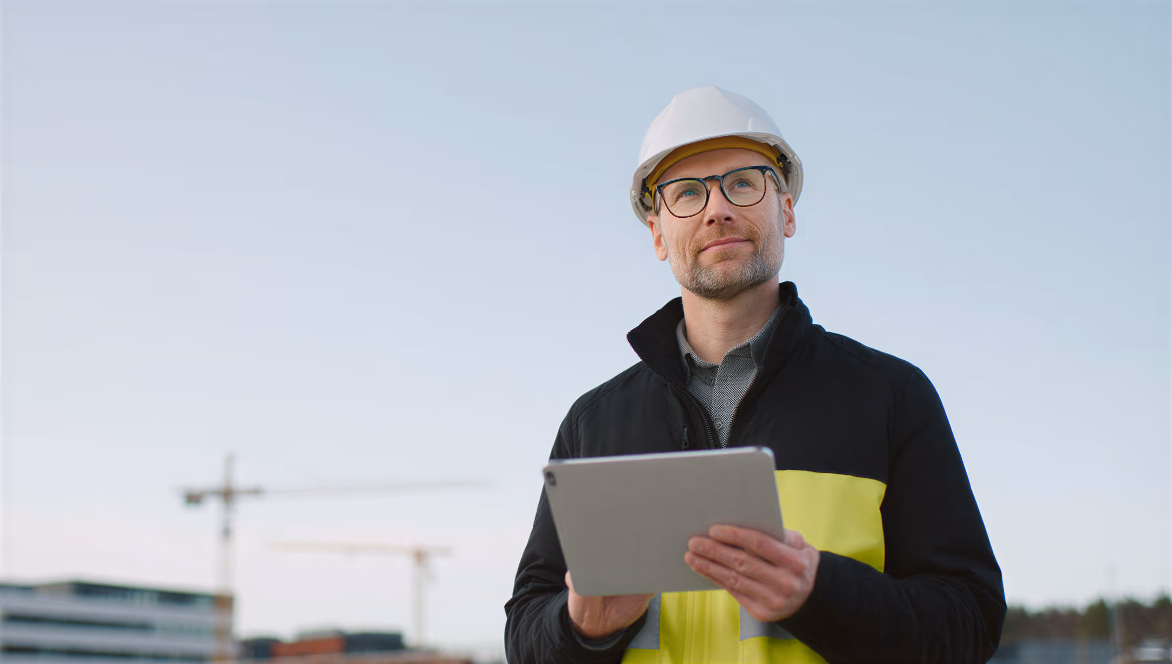 Male construction worker wearing a white hard hat and safety jacket holding a tablet with cranes and buildings in the background.
