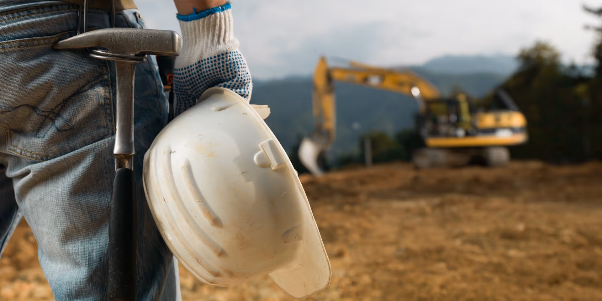 Close-up of construction worker's hand holding a white hard hat with a hammer tucked into jeans pocket, with an excavator blurred in the background.