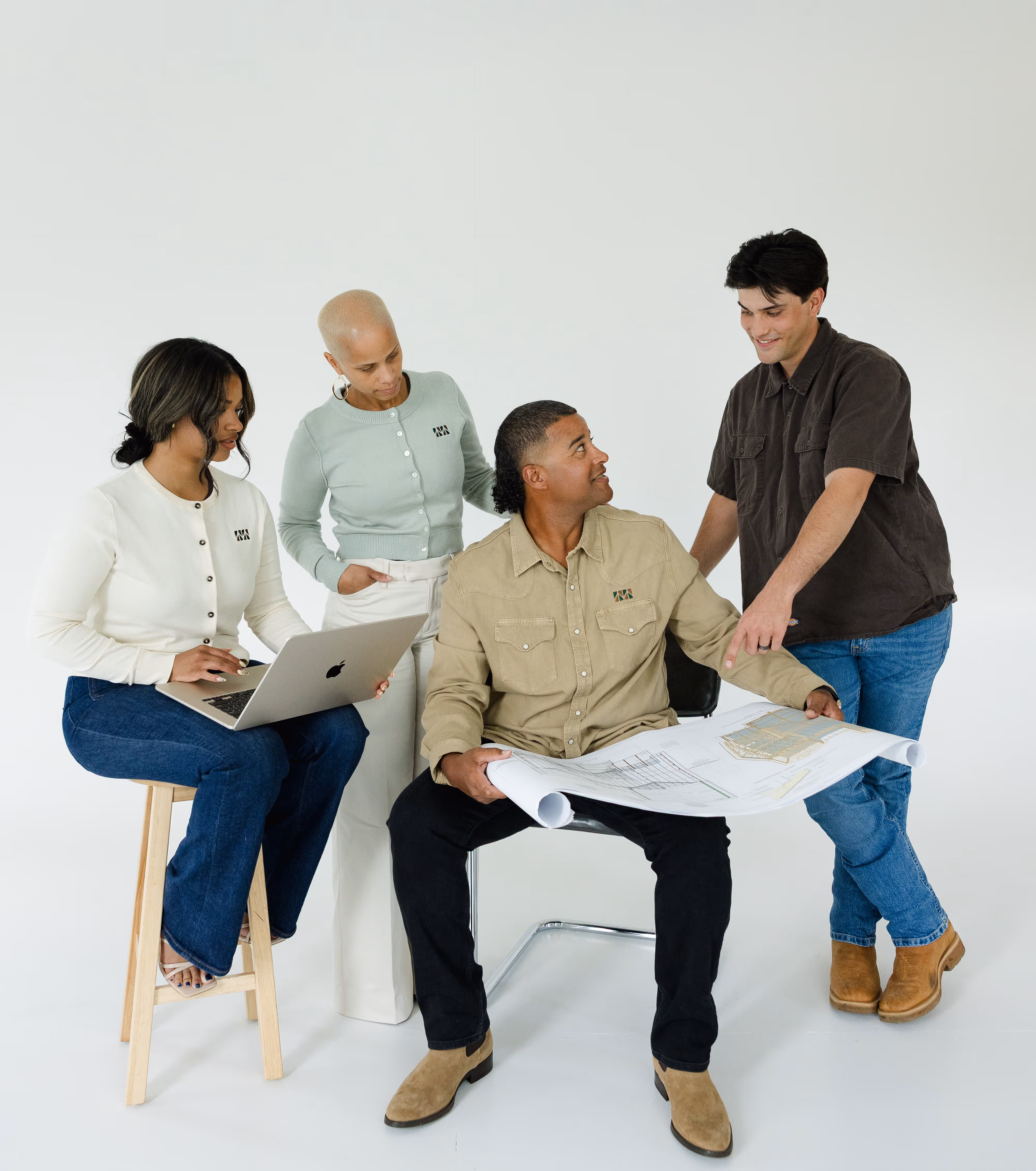 Four people collaborating, one seated with a laptop, one standing with hands in pockets, one seated holding architectural plans, and one standing pointing to the plans.