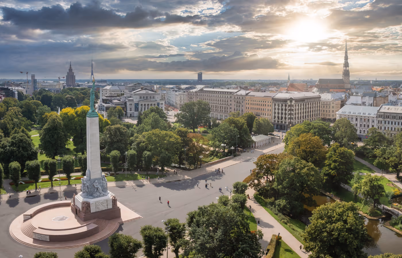 Aerial view of Riga's Freedom Monument surrounded by trees, park pathways, and historic city buildings under a partly cloudy sky.