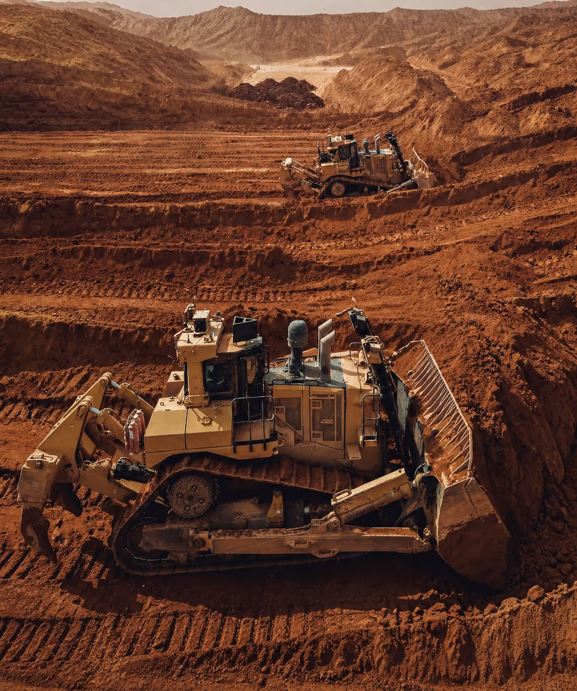 Two large bulldozers moving soil in a terraced, mountainous construction site with brown earth and rugged hills.