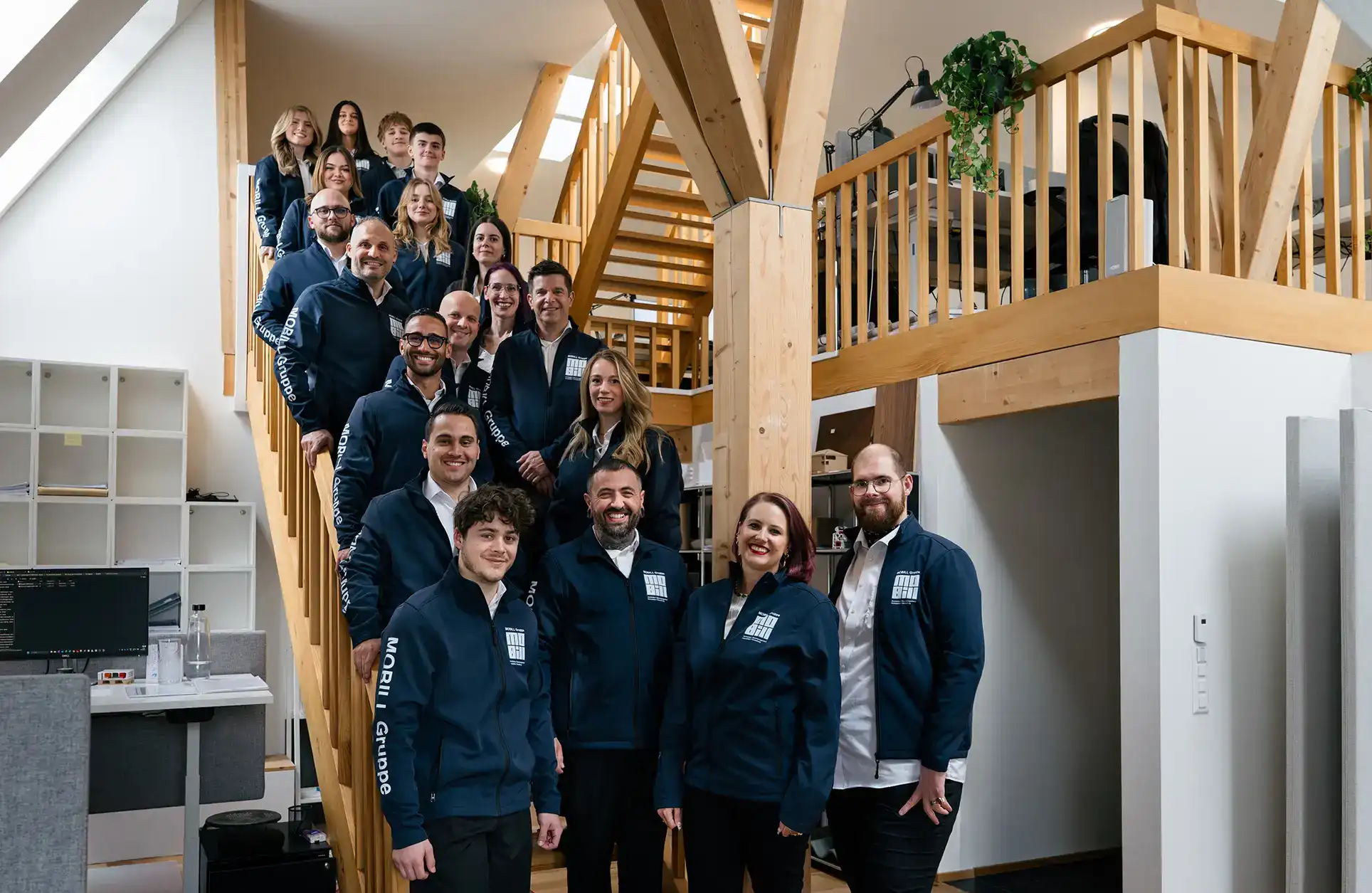 Ein Team von 17 Personen in blauen Jacken steht eng auf einer Holztreppe in einem hellen modernen Büro.