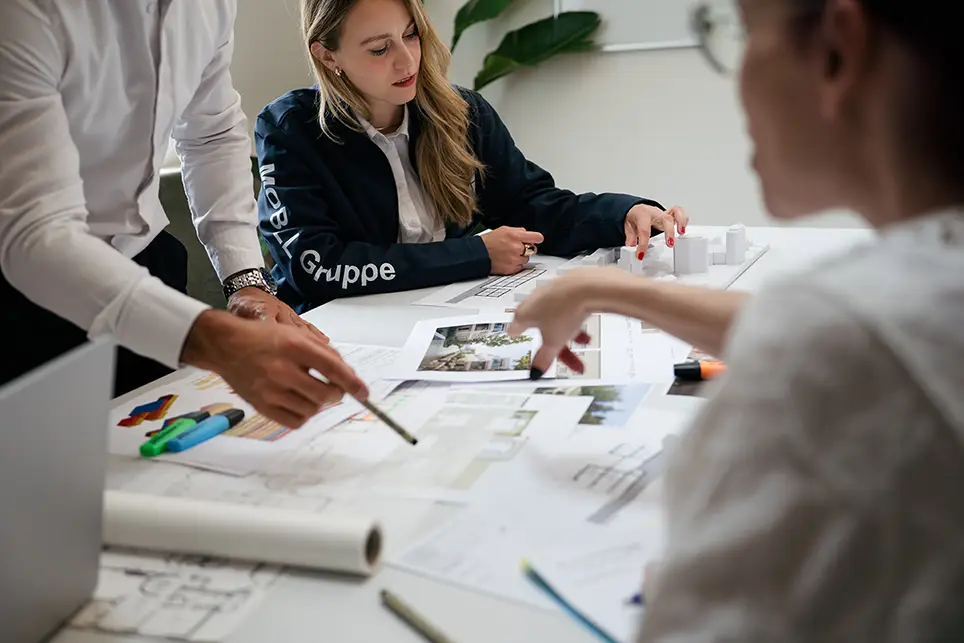 Drei Personen besprechen Architektenpläne und ein Modell auf einem Tisch bei einem Meeting.