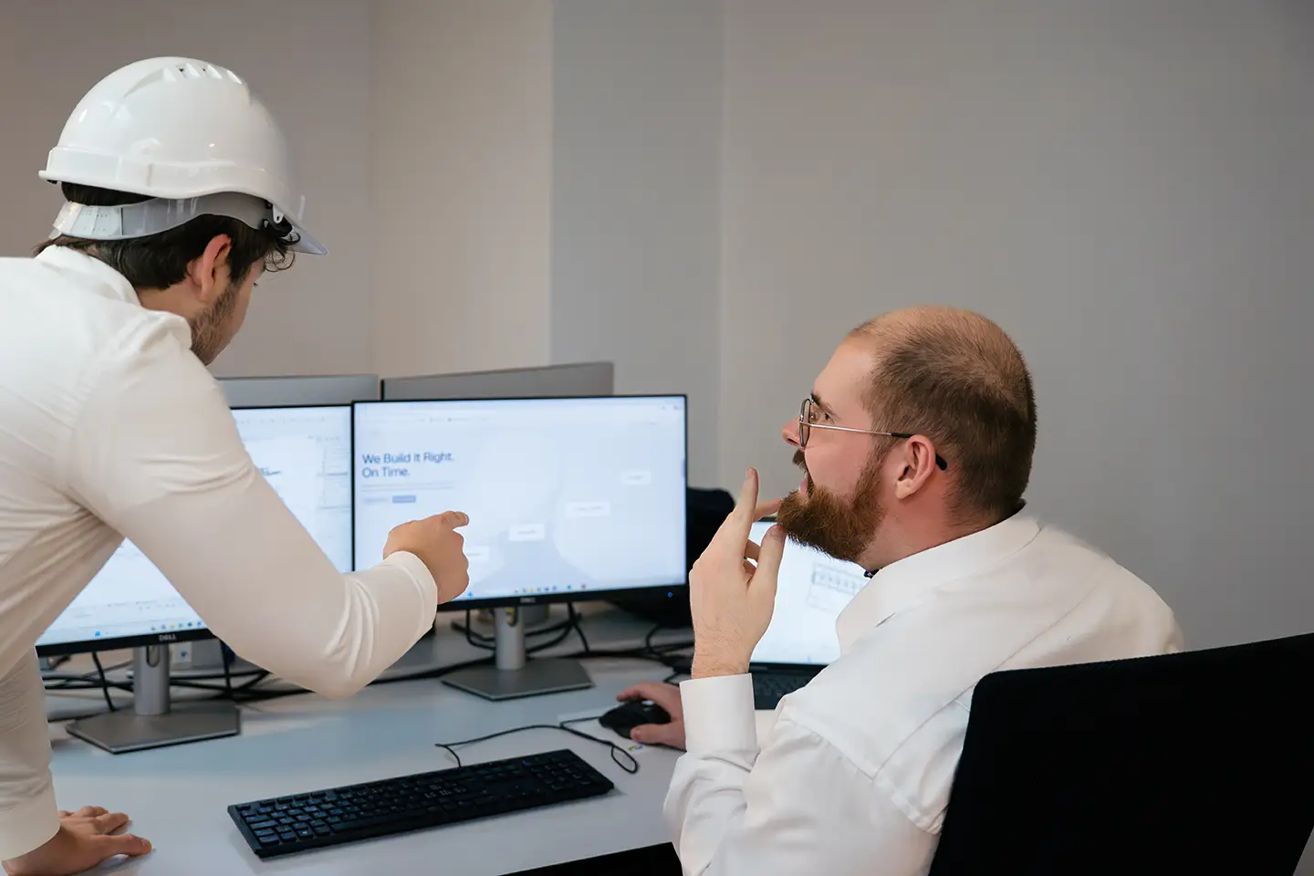Zwei Männer prüfen Baupläne an zwei Desktop-Monitoren, einer trägt einen Schutzhelm.