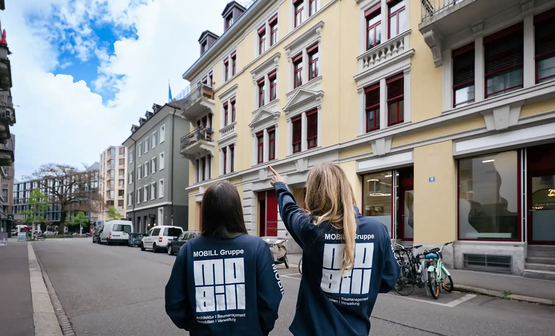 Zwei Frauen mit MOBILL Gruppe Jacken stehen auf einer Stadtstrasse und schauen nach oben auf Gebäude.