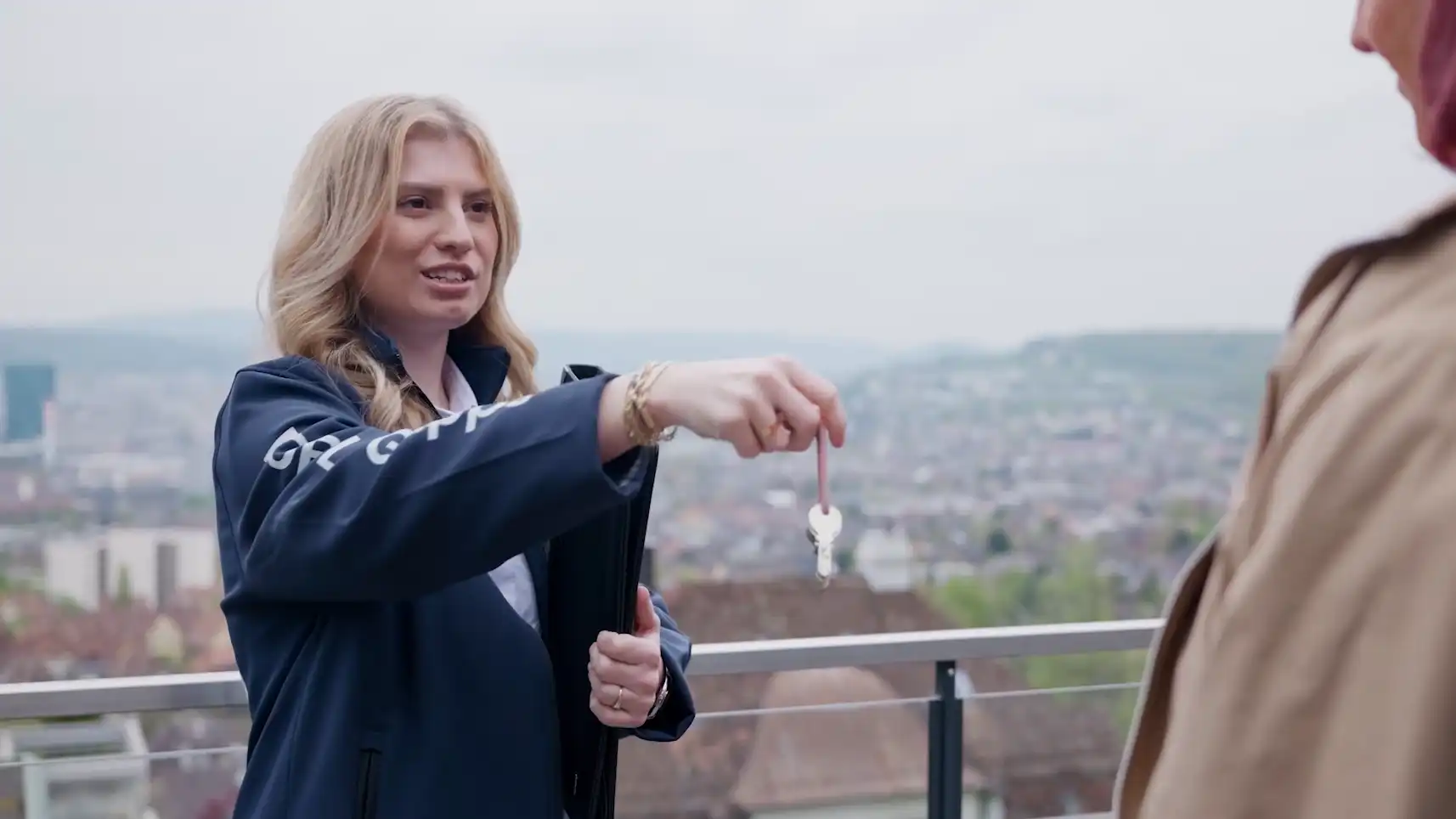 Frau übergibt einem anderen Menschen einen Schlüssel vor einer Stadtlandschaft.