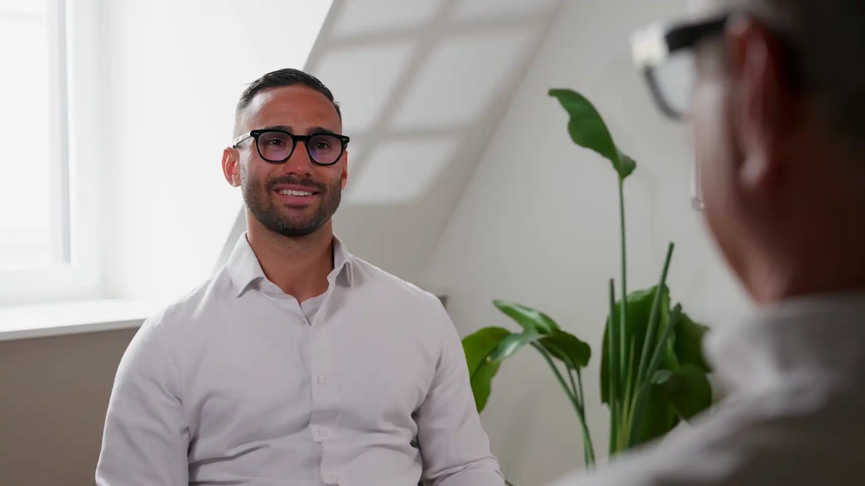 Zwei Männer in Hemden führen ein Gespräch in einem hellen Büro mit grüner Pflanze im Hintergrund.