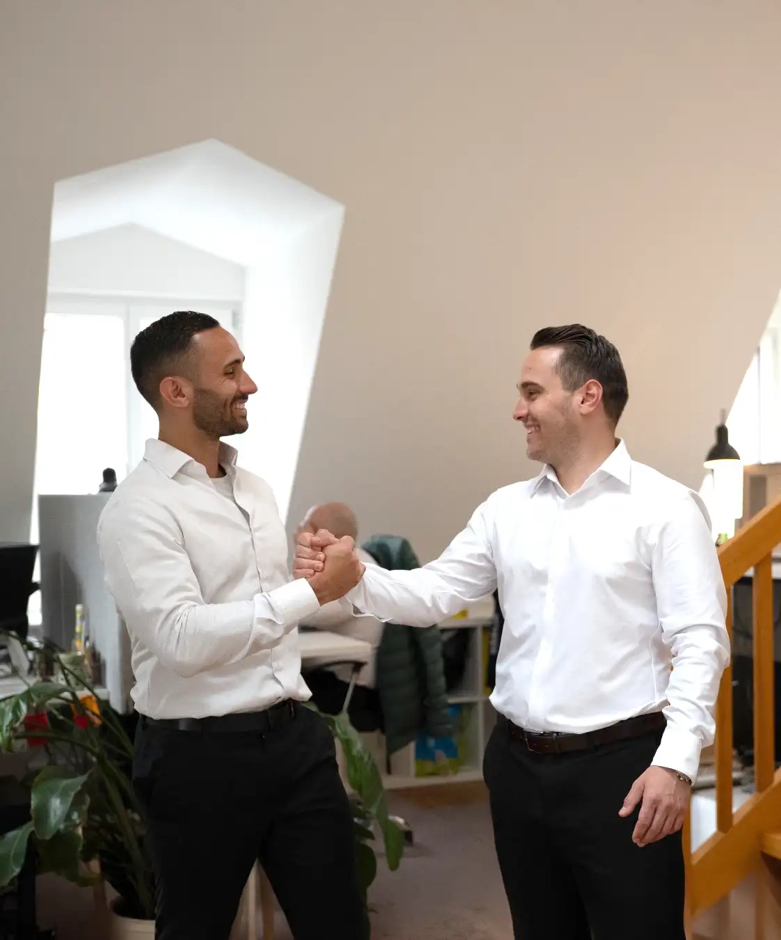 Zwei Männer in weißen Hemden schütteln sich in einem Büro die Hände und lächeln sich an.