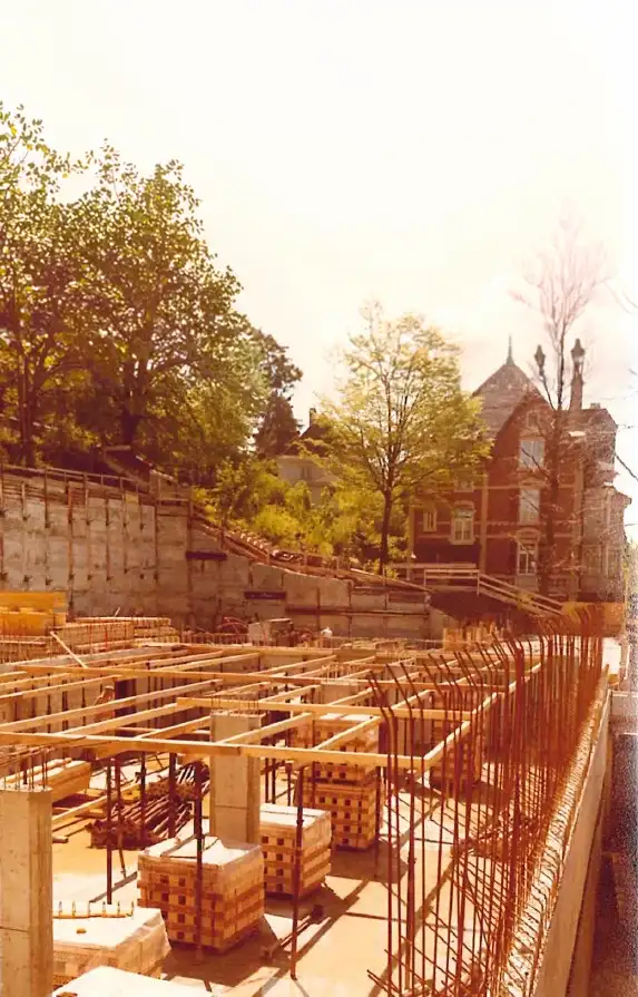 Baustelle mit Stahlbewehrungen und Holzrahmen neben einem roten Backsteingebäude und Bäumen.