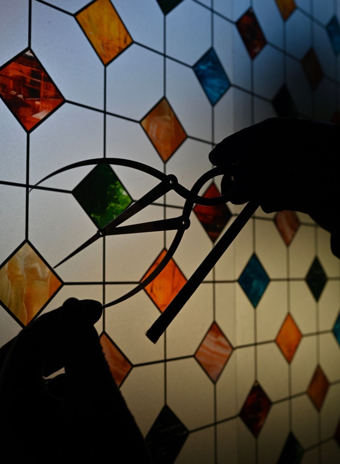 Silhouetted hands holding a divider and pencil in front of a stained glass window with colorful diamond patterns.