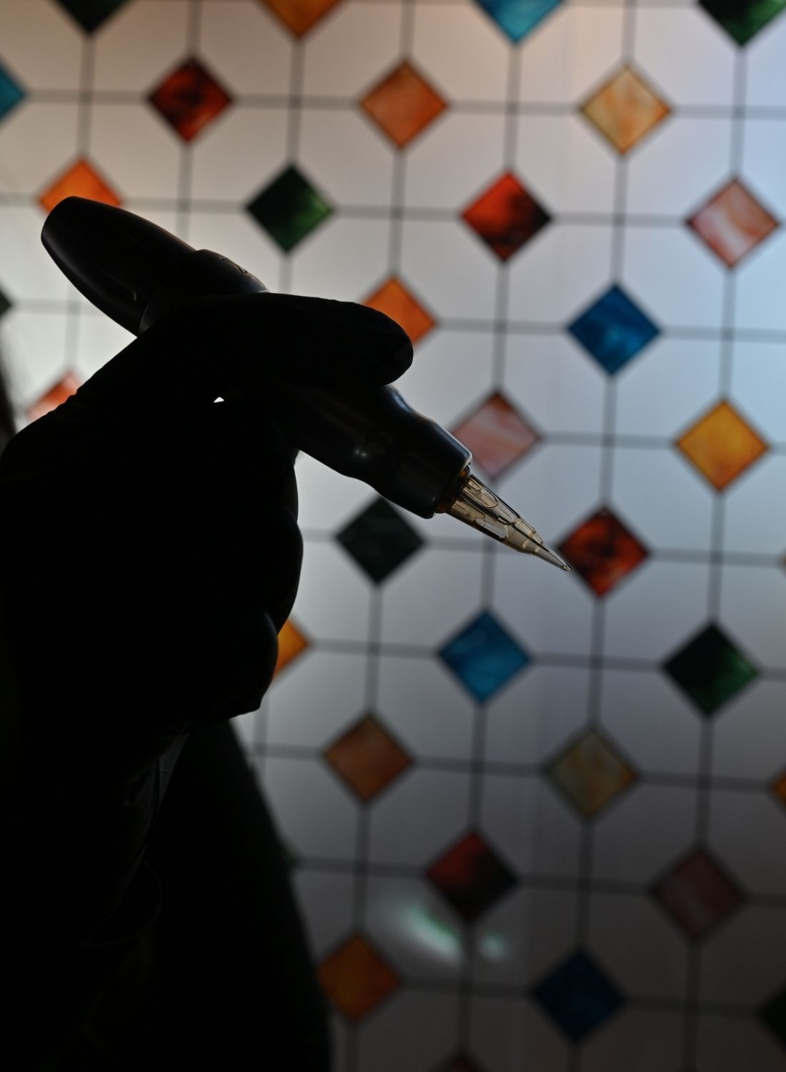 Silhouette of a gloved hand holding a tattoo machine against a colorful tiled background.