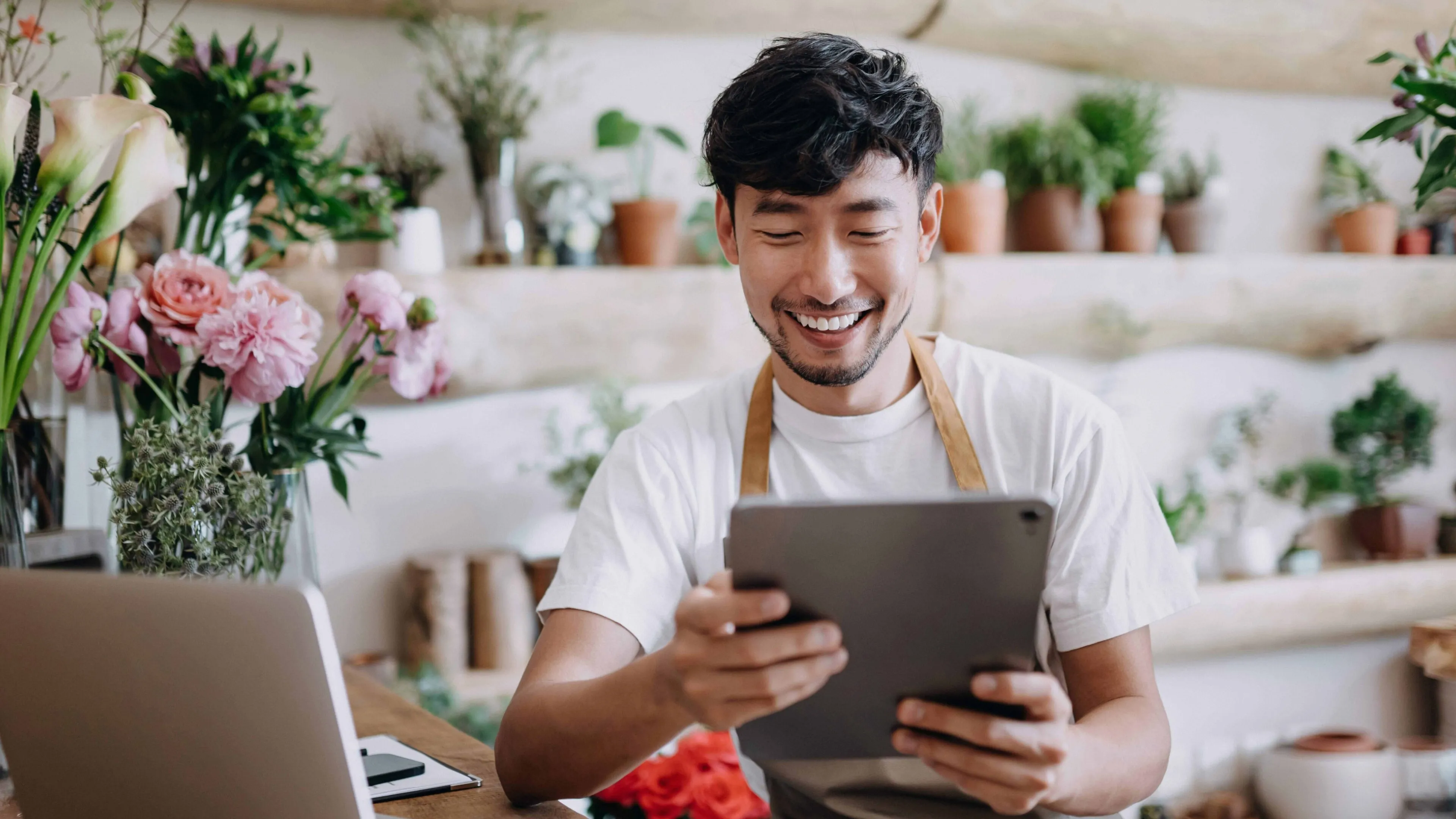 Smiling florist wearing a white shirt and brown apron sits at a wooden table surrounded by plants and flowers, holding a tablet. A laptop and documents are on the table, with shelves of greenery in the background