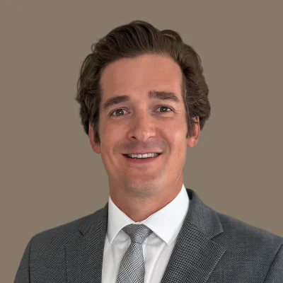 Smiling man with brown hair wearing a gray suit, white shirt, and patterned tie on a brown background.
