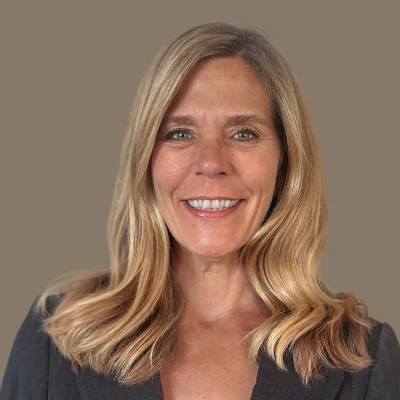 Smiling woman with long blonde hair wearing a dark blazer against a plain background.