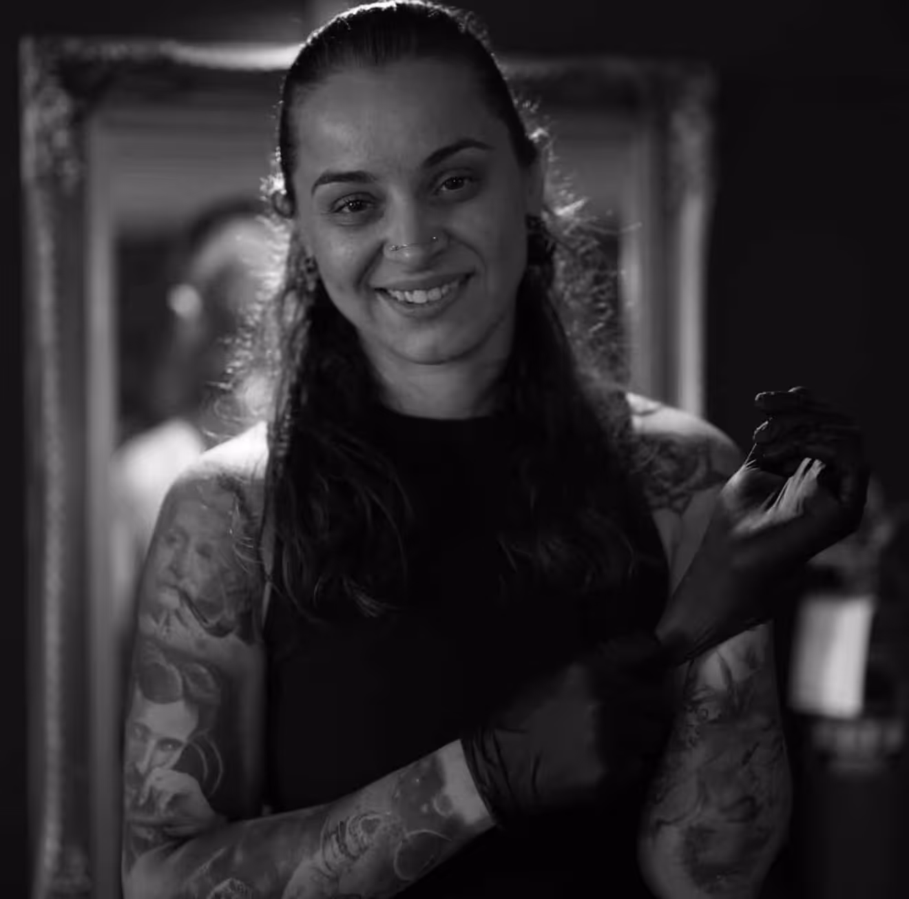Smiling woman with multiple tattoos on her arms putting on a black glove in a dimly lit room.