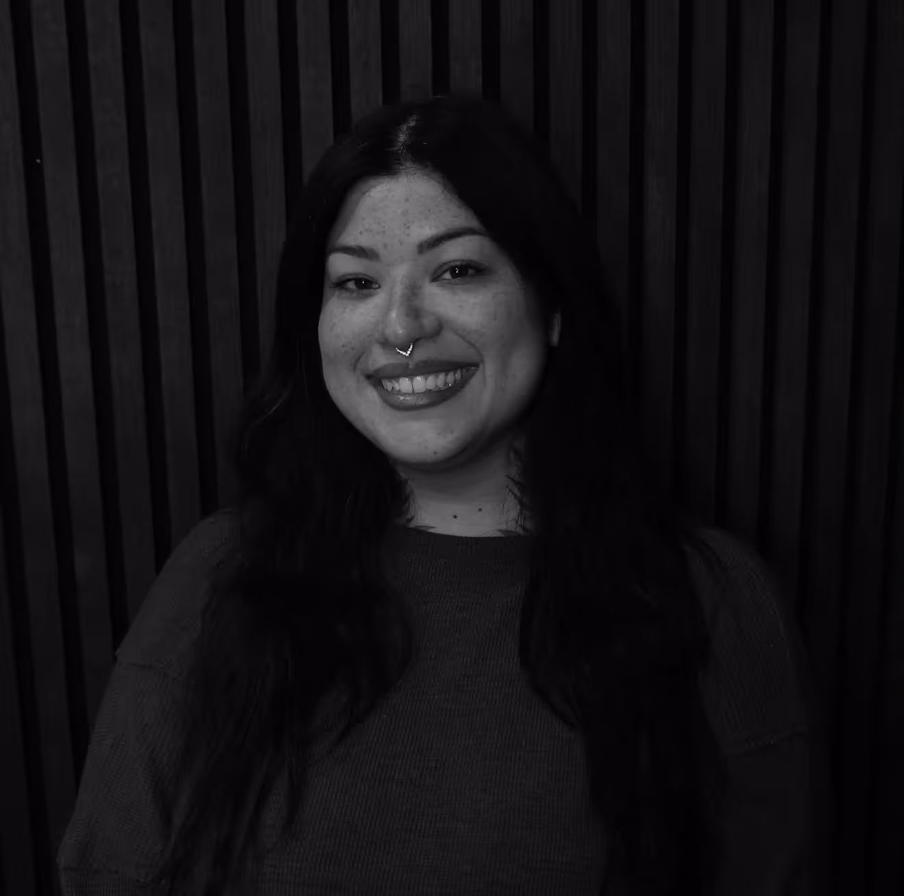 Smiling woman with long dark hair and a septum nose ring against a dark vertical striped background.
