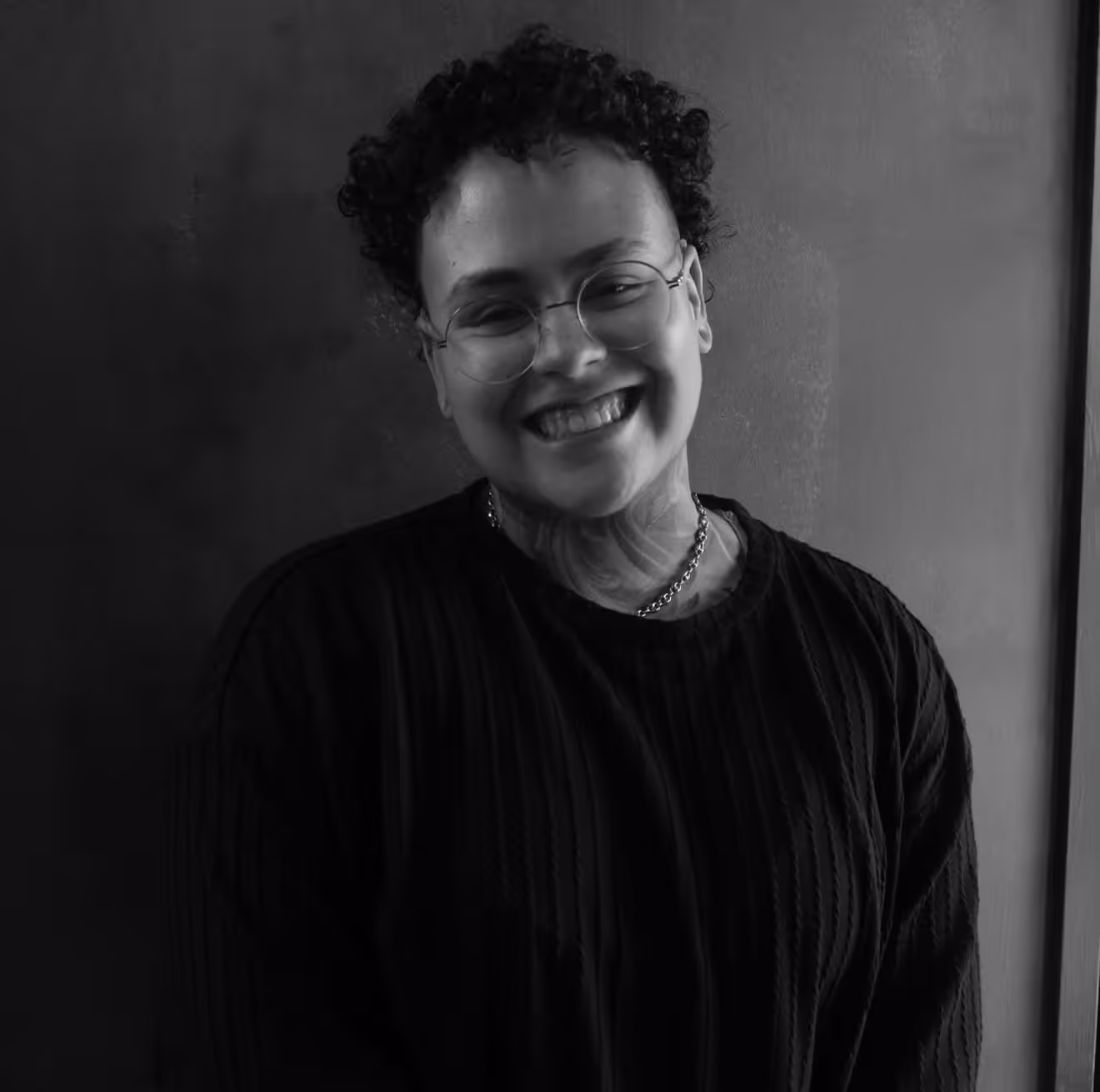 Smiling person with curly hair, glasses, a chain necklace, and a dark ribbed sweater standing against a plain background.
