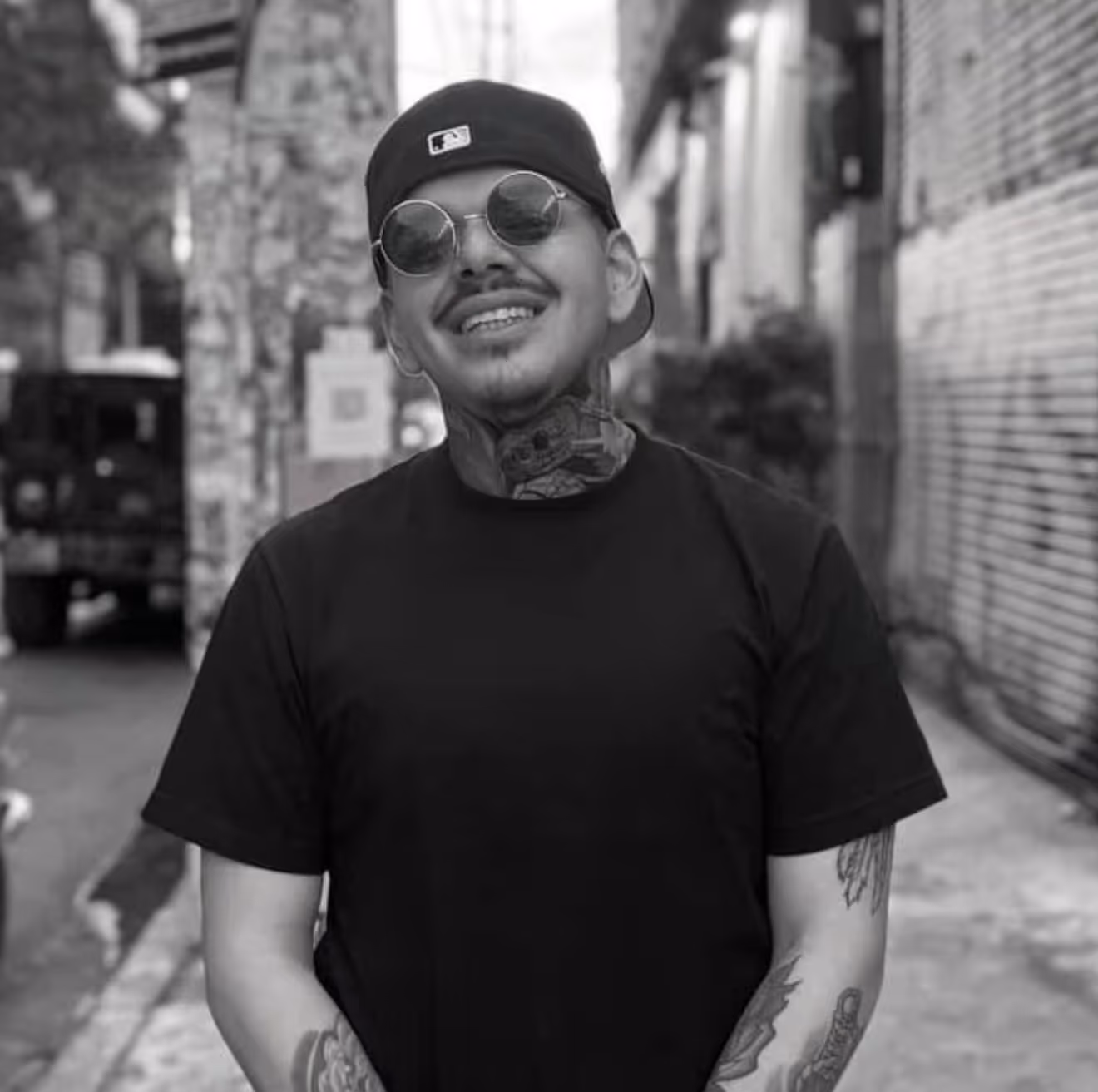 Smiling man with tattoos on neck and arms wearing round sunglasses, a black backwards cap, and a black t-shirt in an urban alley.