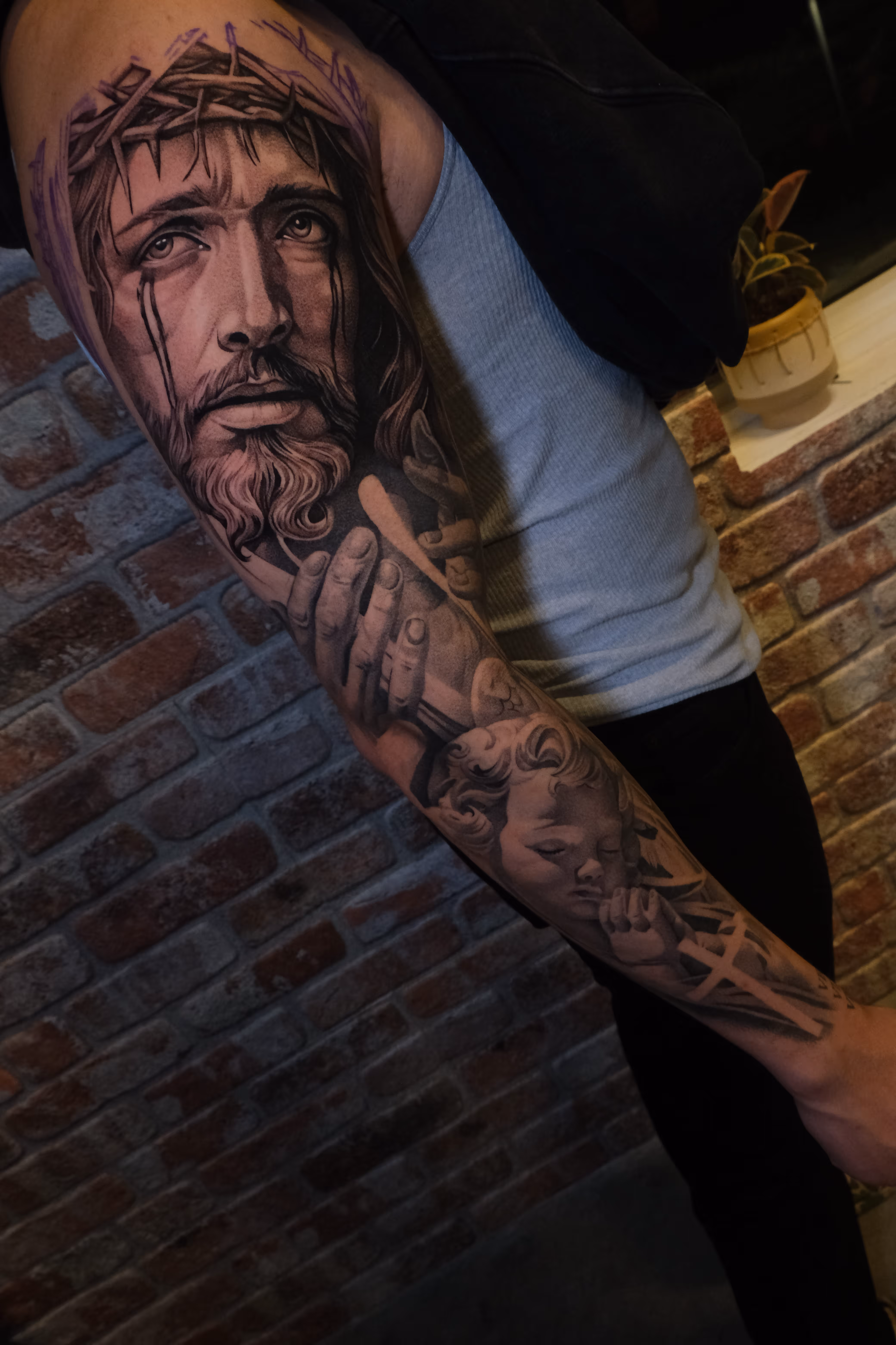 Tattoo sleeve featuring a realistic face of Jesus wearing a crown of thorns with tears and a praying cherub below on a person's arm.