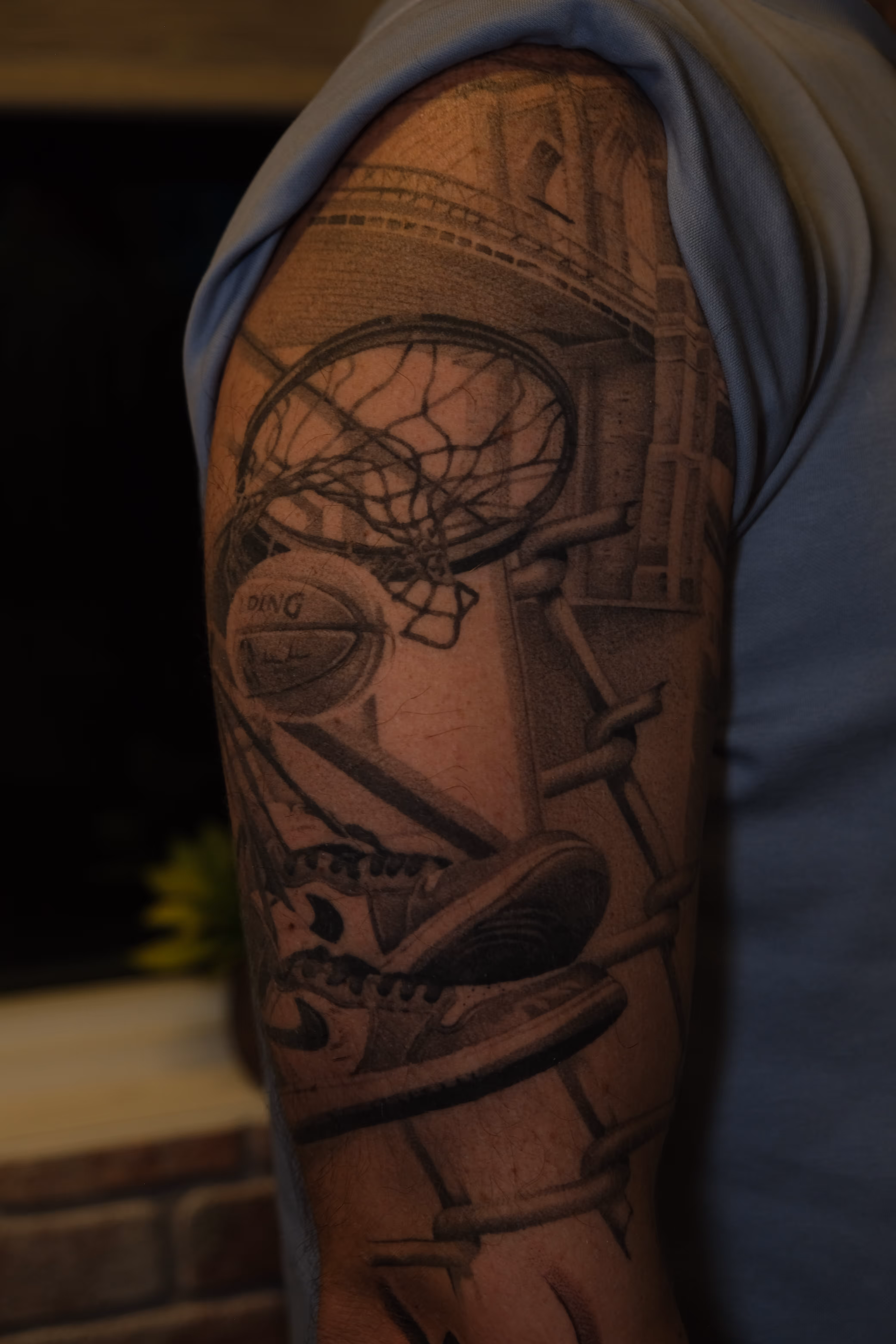 Black and gray tattoo on upper arm depicting a basketball hoop with net, a basketball going through the net, and a pair of Nike sneakers.
