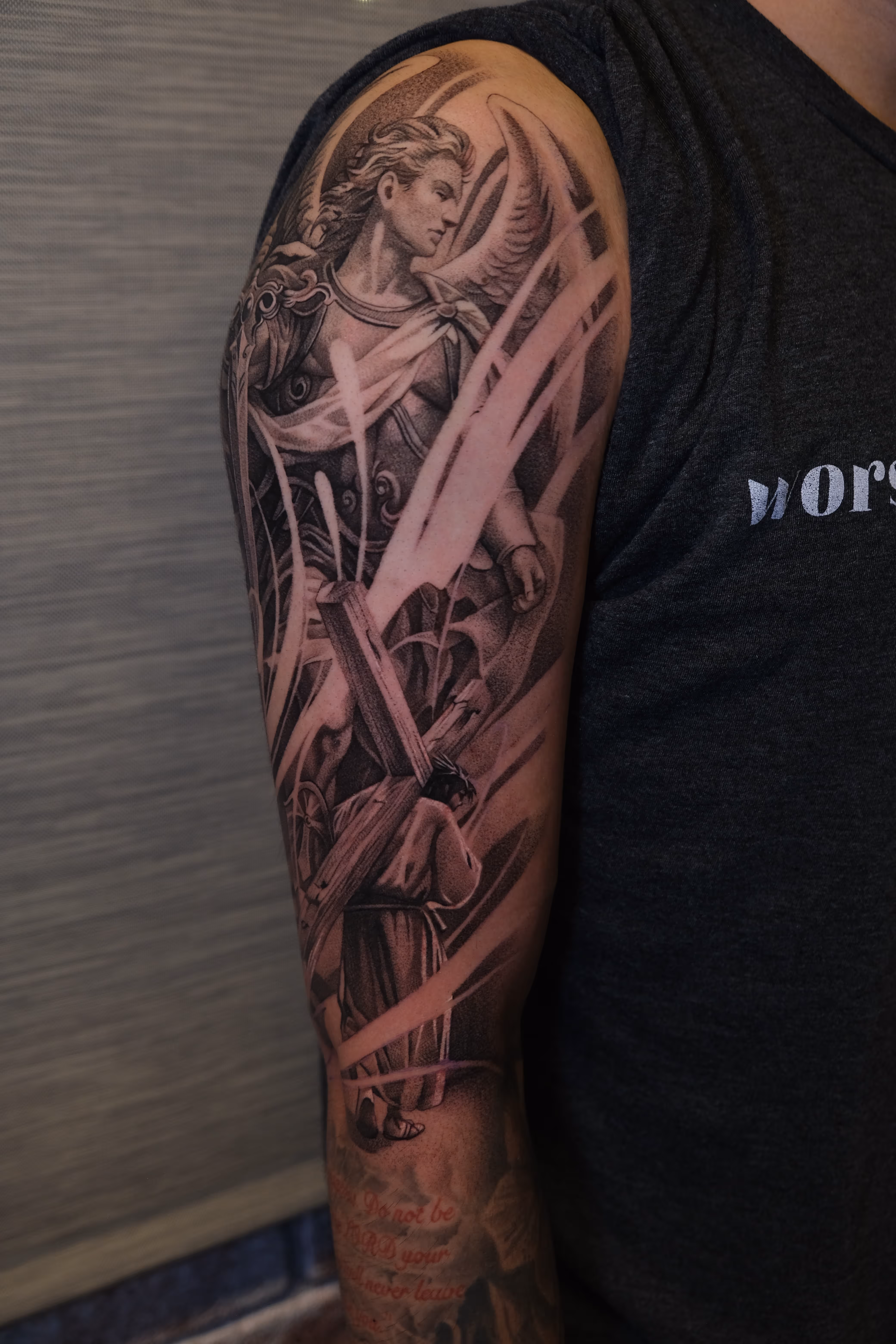 Black and gray tattoo of a winged angel and a man carrying a wooden cross on a person's upper arm.