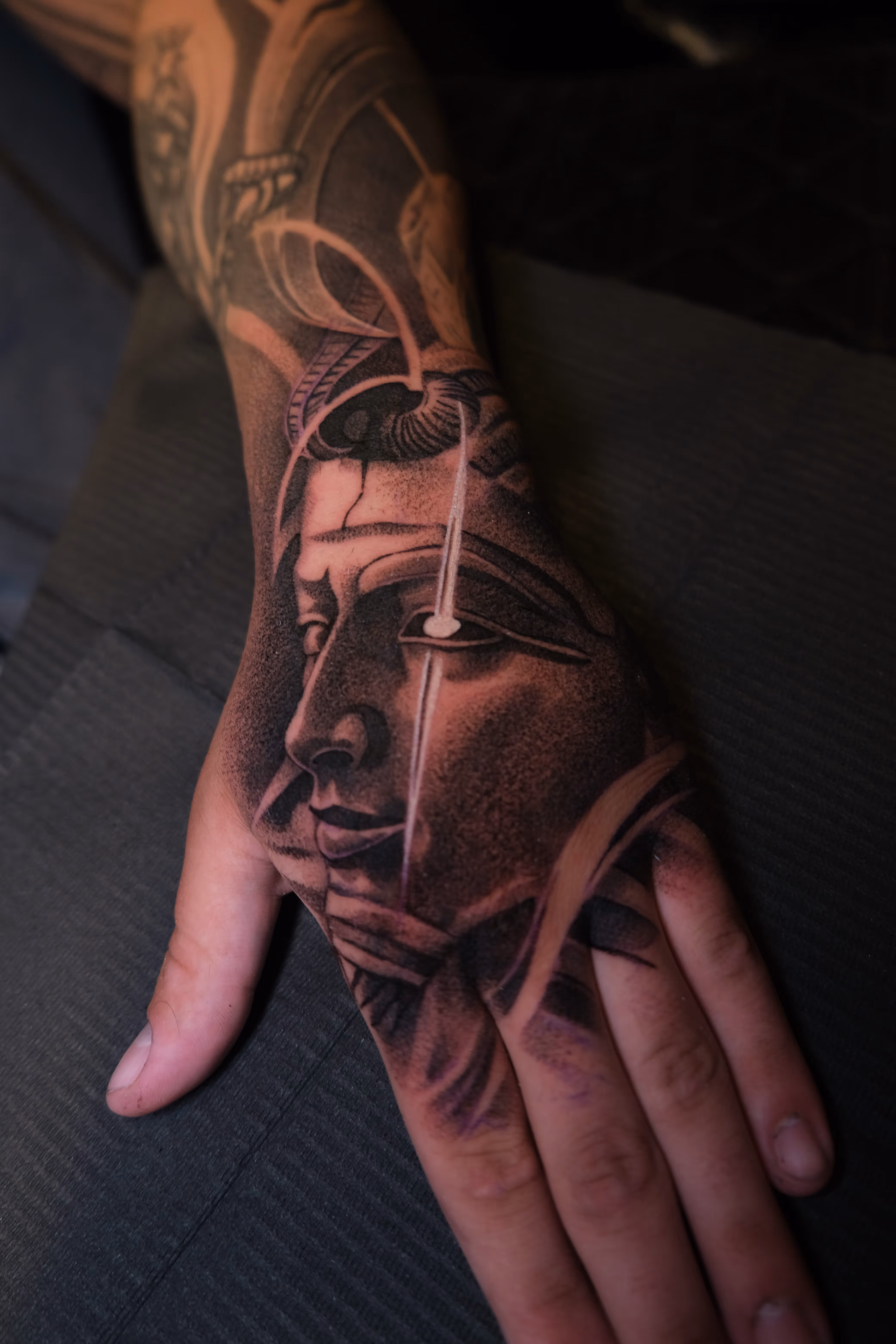 Tattoo on hand featuring a detailed, shaded face with an eye design and white highlights.