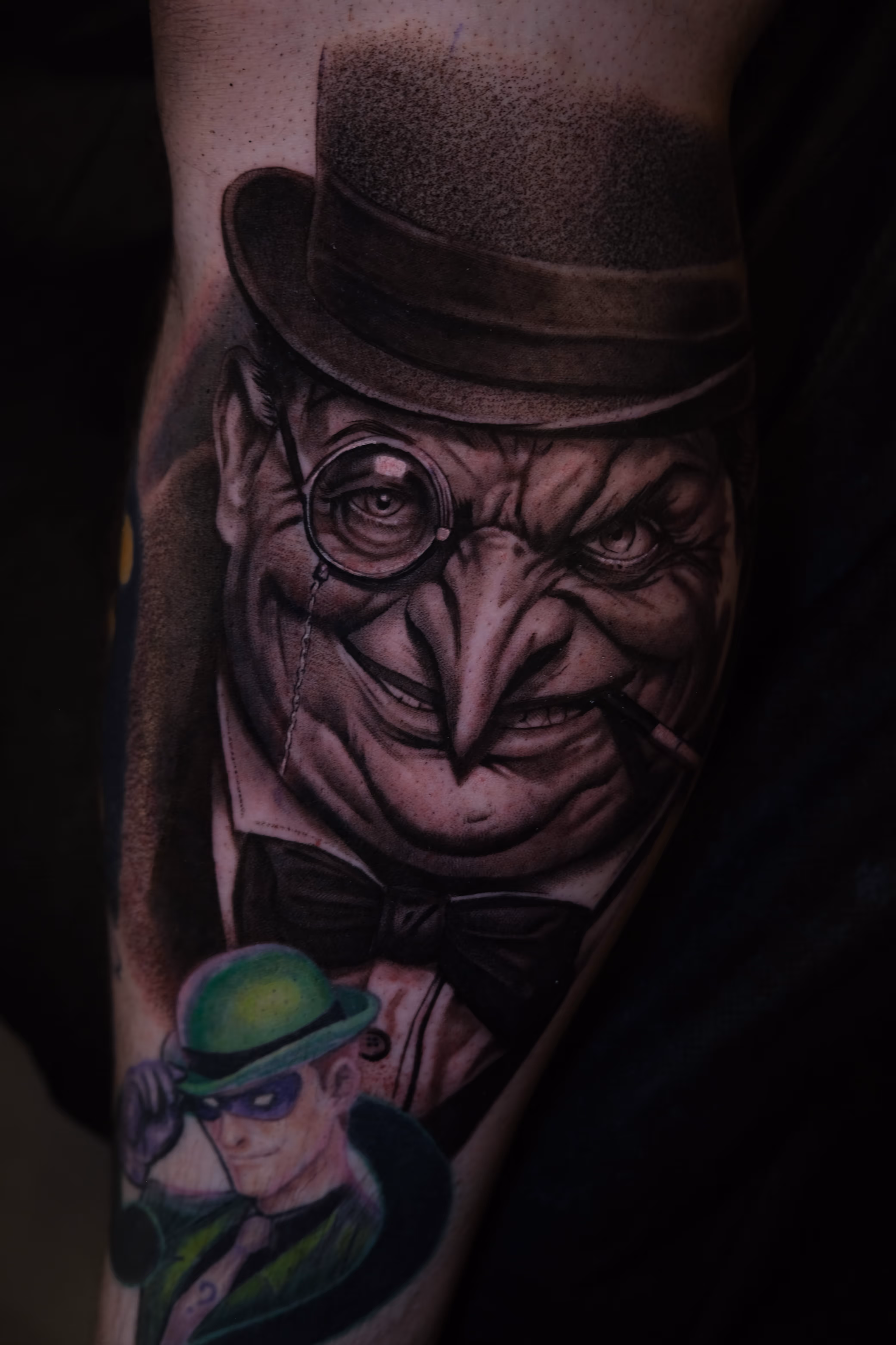 Tattoo of the comic book villain Penguin with a long nose, monocle, top hat, and bow tie, accompanied by a smaller tattoo of the Riddler in a green bowler hat and mask.
