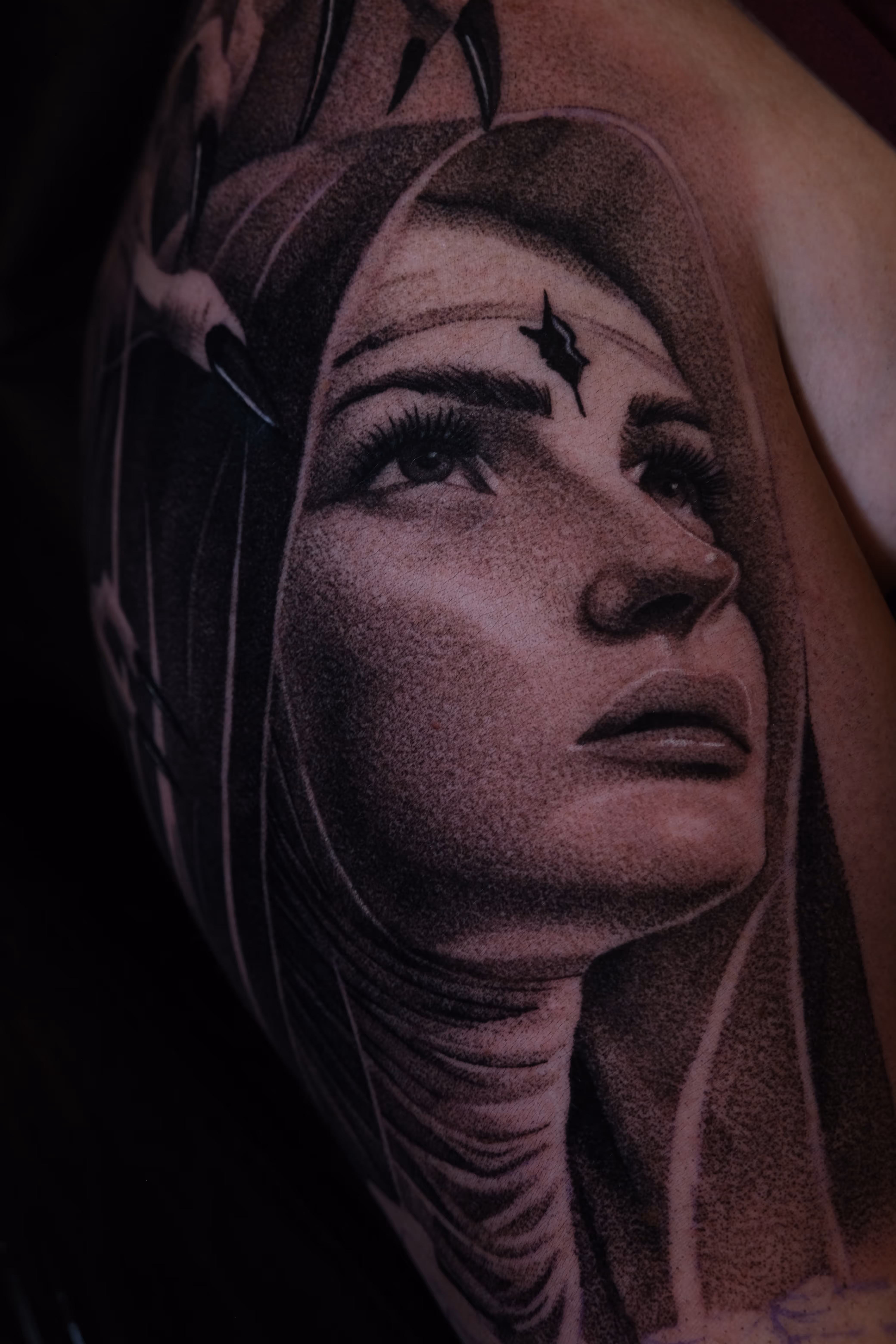 Black and gray tattoo of a woman’s face in a hood looking upward, with detailed shading and a star-shaped mark on her forehead.