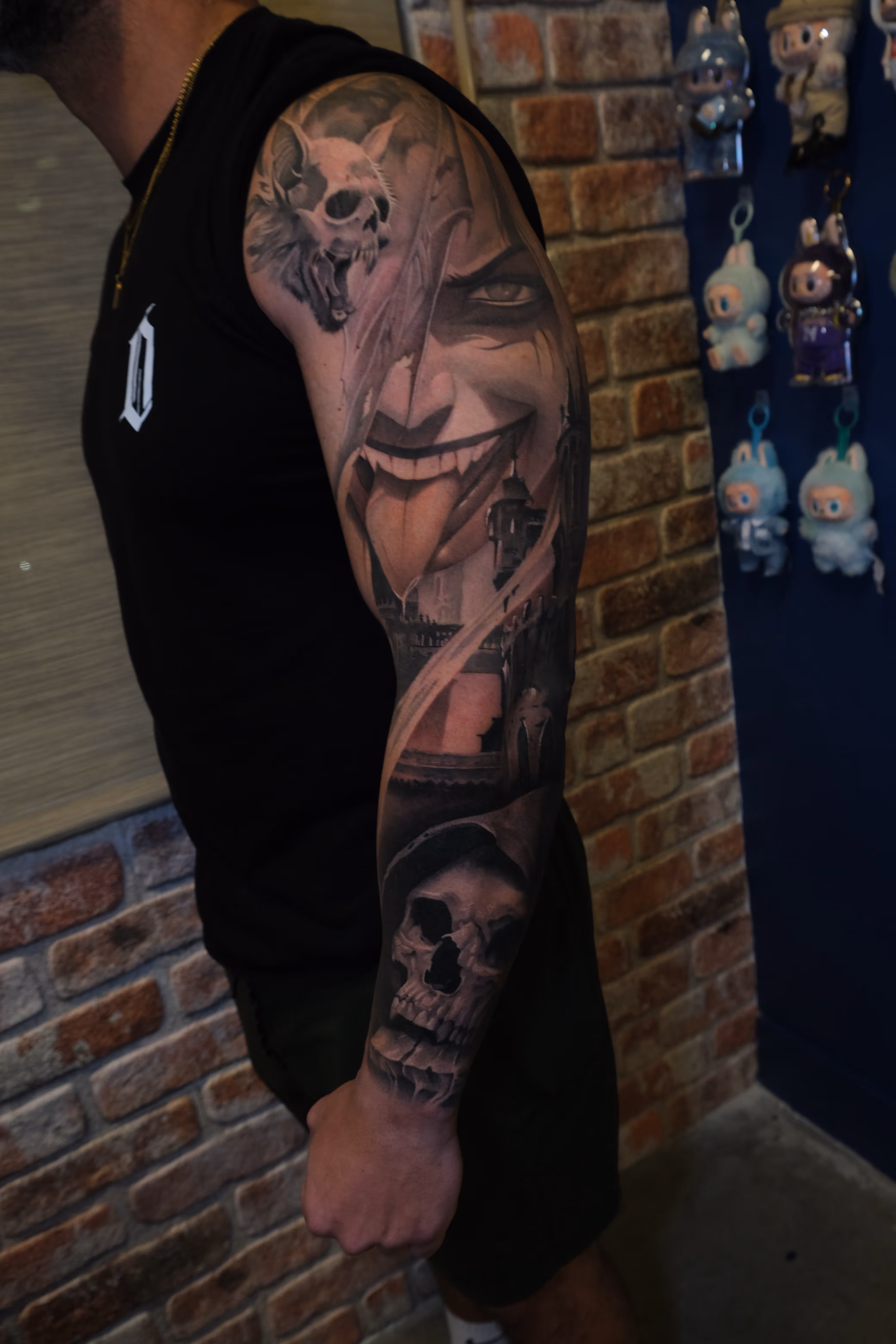 Detailed full sleeve tattoo of a large grinning face with a protruding tongue, a snarling wolf skull, a castle, and a grim reaper skull on a person's arm.