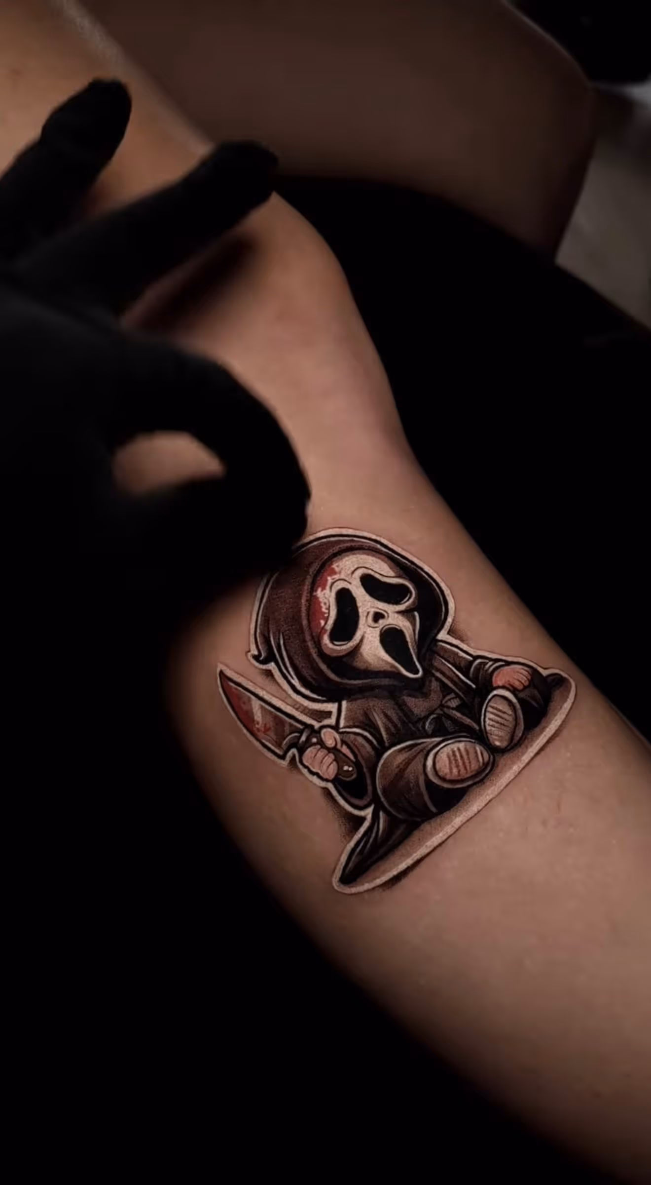 Tattoo of a cartoon Grim Reaper with a bloody knife on a person's forearm with a gloved hand nearby.