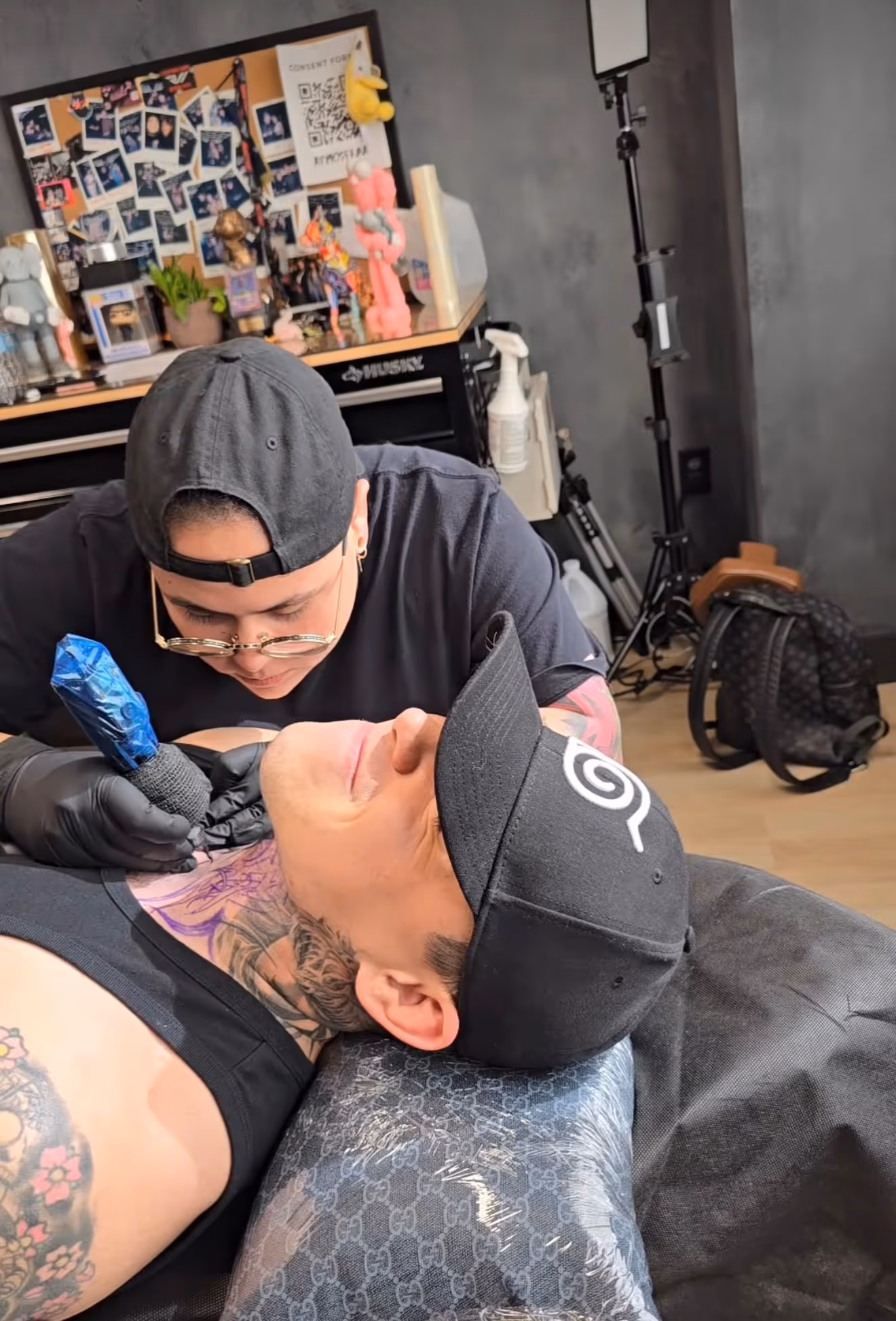 Tattoo artist wearing glasses and a black cap tattooing a man's neck with a black tank top and a black cap in a tattoo studio.
