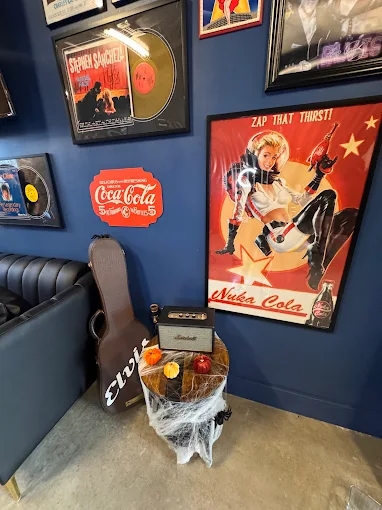Corner of a room with blue walls decorated with vintage music and Coca-Cola posters, a black guitar case, a small amplifier on a stool covered with fake spider webs, and small pumpkins.