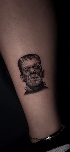 Detailed full sleeve tattoo of a large grinning face with a protruding tongue, a snarling wolf skull, a castle, and a grim reaper skull on a person's arm.
