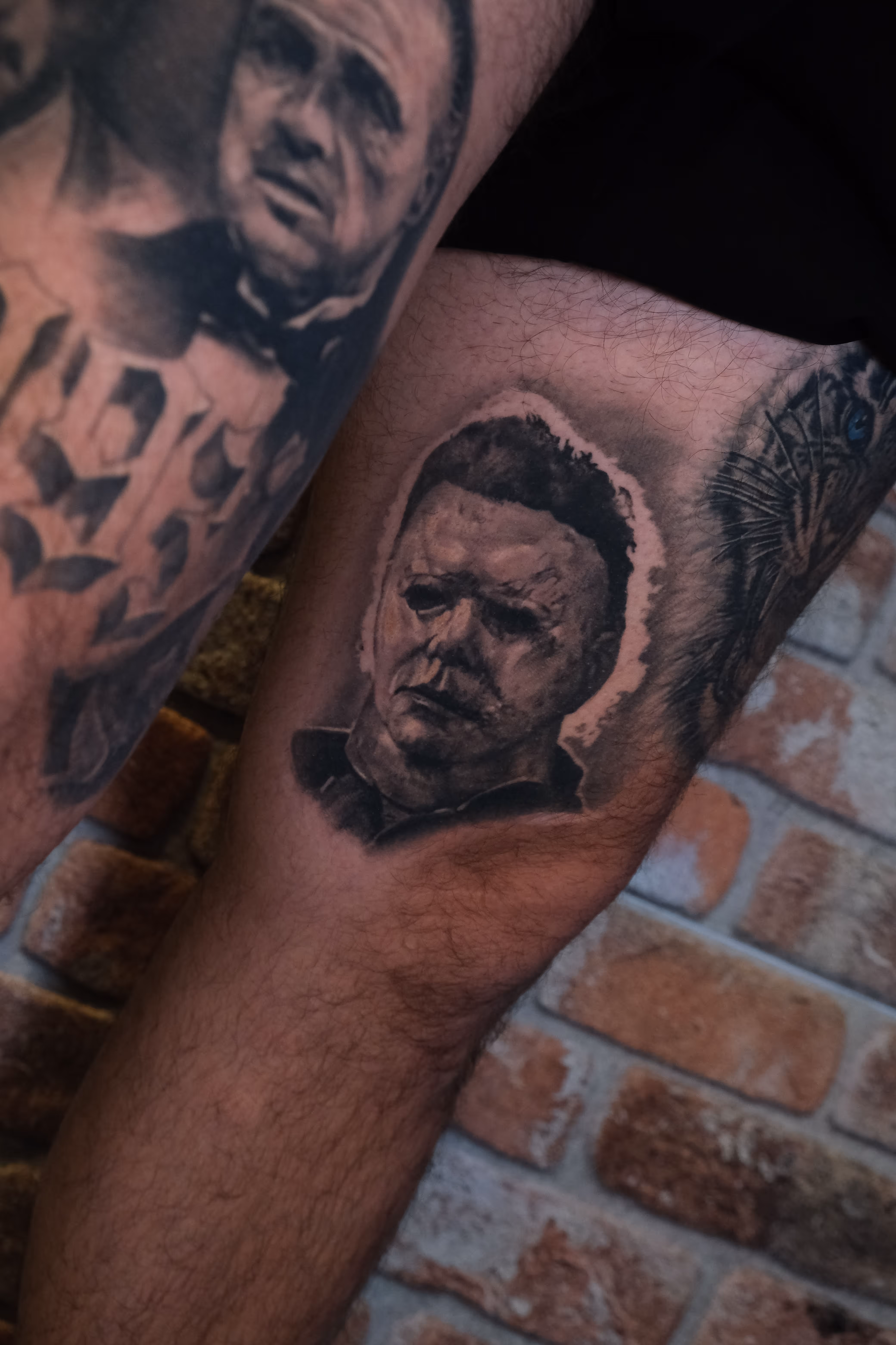 Detailed full sleeve tattoo of a large grinning face with a protruding tongue, a snarling wolf skull, a castle, and a grim reaper skull on a person's arm.