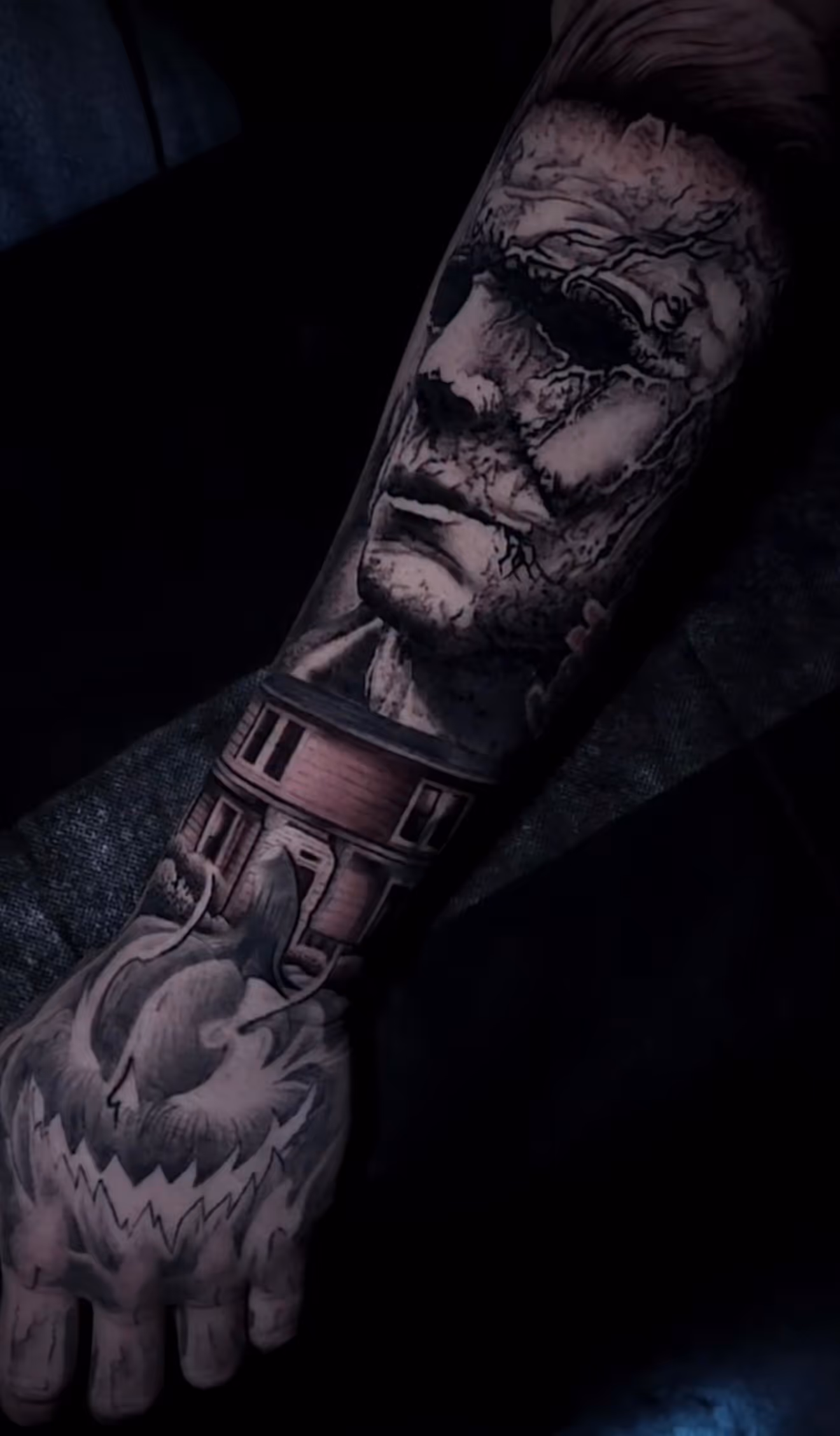 Detailed full sleeve tattoo of a large grinning face with a protruding tongue, a snarling wolf skull, a castle, and a grim reaper skull on a person's arm.