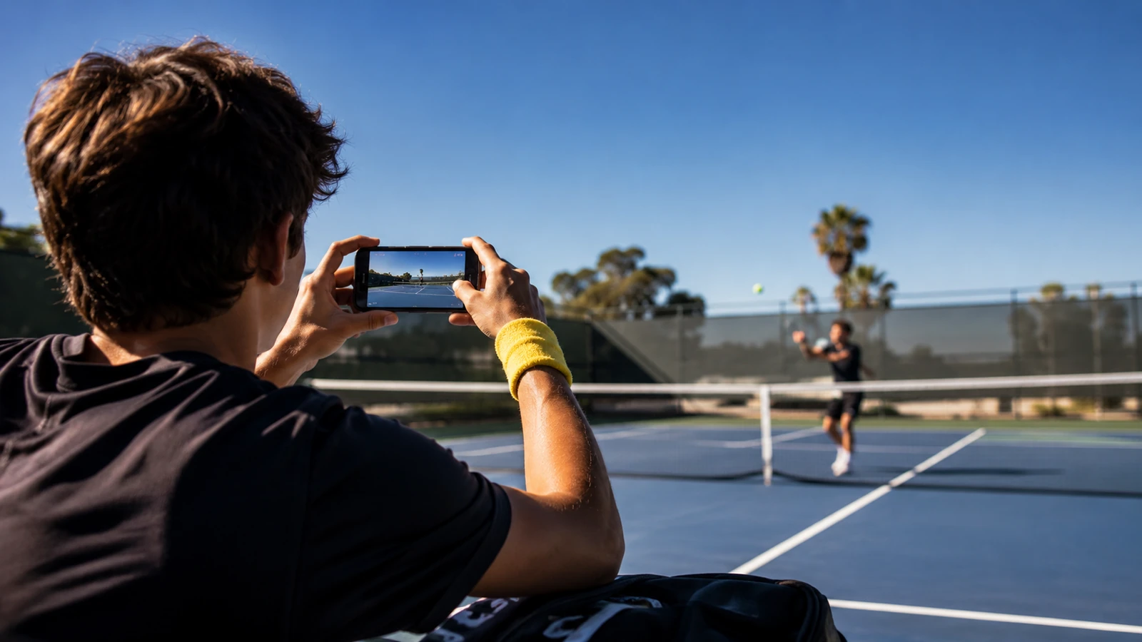 Junior tennis player filming a practice clip on a phone on a sunlit court