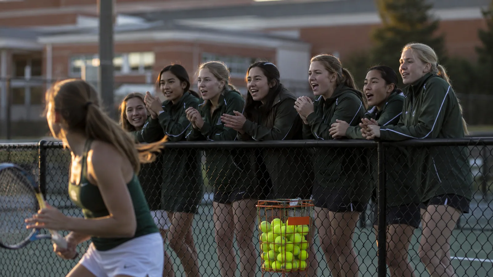 High school tennis teammates cheering courtside behind the fence during a match