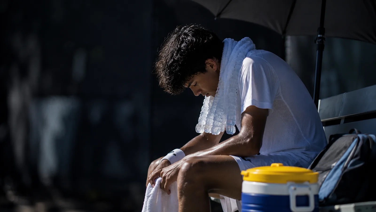 Tennis player cooling down with an ice collar under an umbrella during a changeover in intense sun