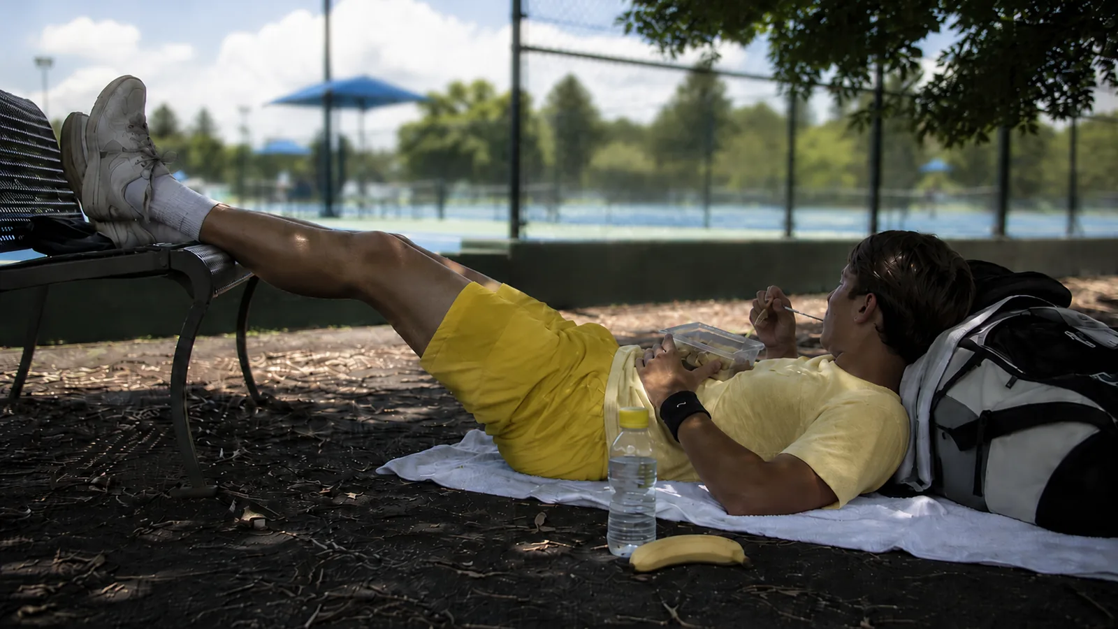 Tennis player resting in the shade with legs elevated and a recovery snack between tournament matches