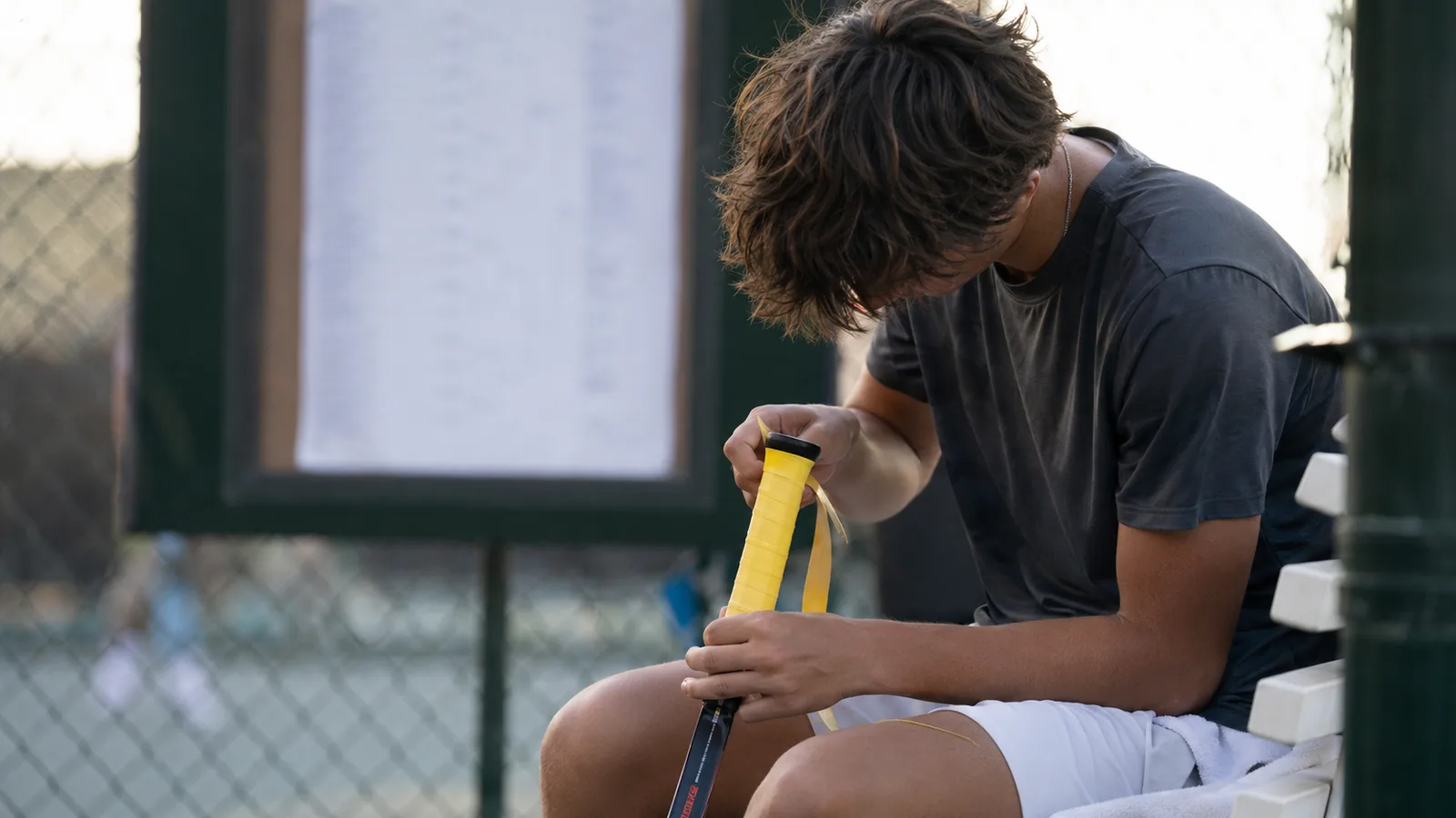 Tennis player re-gripping a racquet on a bench with a tournament draw board behind