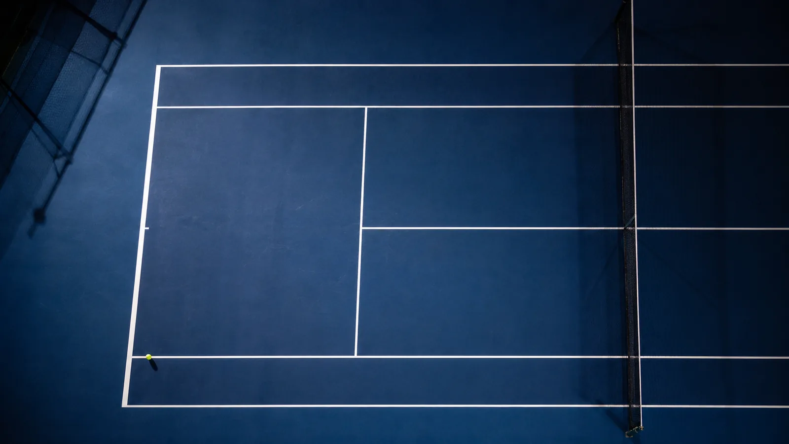 Overhead night view of an empty blue hard court with strong white line geometry and a single ball near the corner