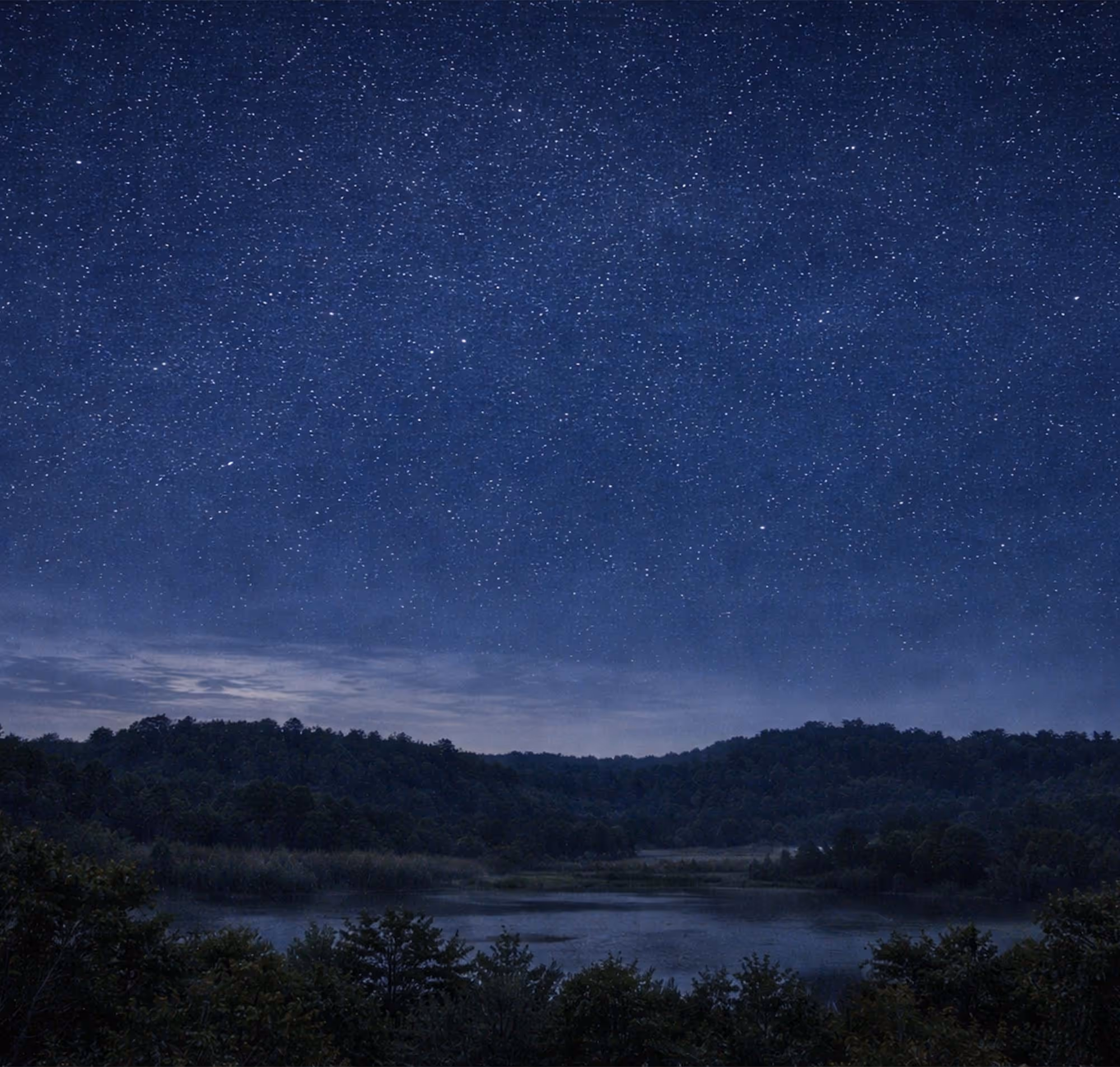 Cielo estrellado sobre un paisaje nocturno con colinas y un lago rodeado de árboles.