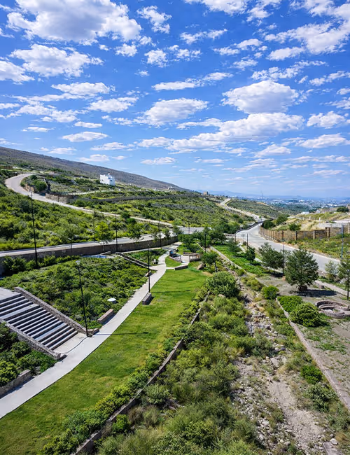 Parque con caminos pavimentados, césped verde y arbustos, con colinas en el fondo bajo un cielo azul con nubes blancas.