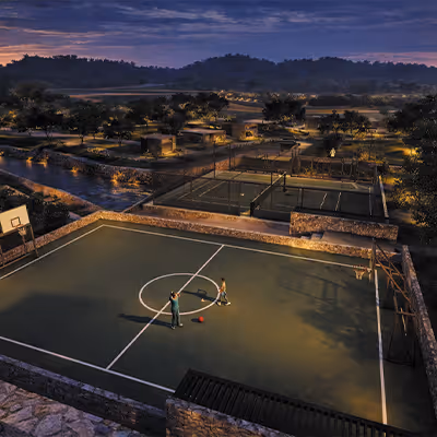 Cancha de baloncesto al aire libre iluminada durante el atardecer con dos personas jugando y un paisaje de colinas en el fondo.