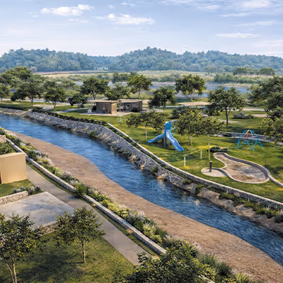 Parque con río, área de juegos infantiles con resbaladilla y columpios, rodeado de árboles y senderos peatonales.