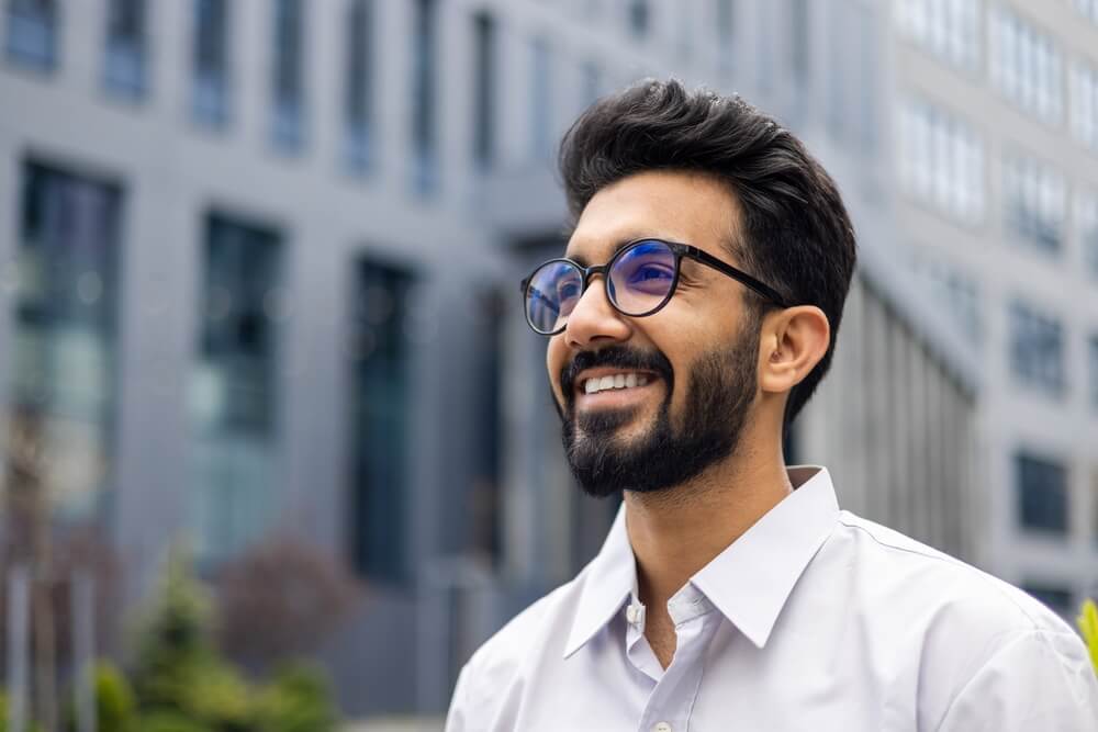Optimistic young adult with beard, glasses, and a white shirt enjoying a sunny day in the city. Casual urban professional.